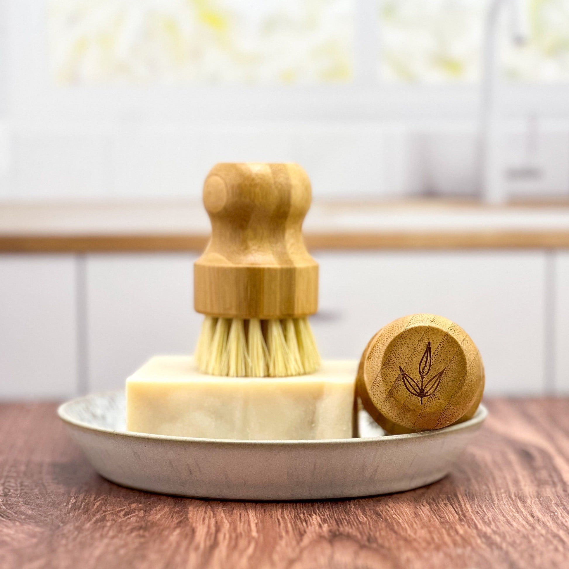 The MamaSuds Bamboo Pot Brush with natural sisal bristles rests on soap in a ceramic dish on a wooden countertop, its matching wooden lid with a leaf design nearby. A softly blurred kitchen forms the backdrop.