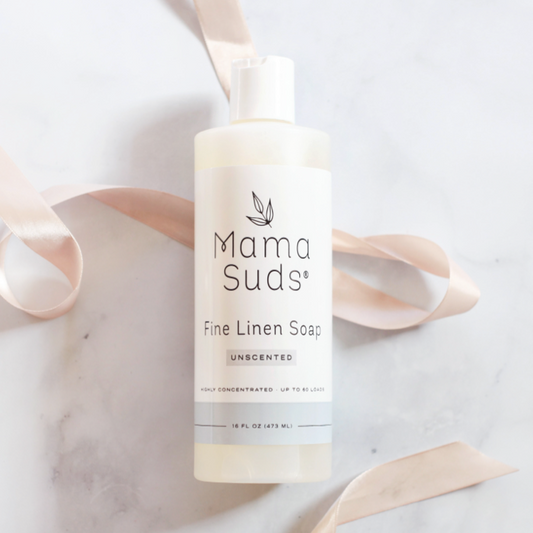 A 16 fl oz (473 mL) bottle of MamaSuds Fine Linen Soap sits on a white surface, adorned with a light pink ribbon. This non-toxic, highly concentrated linen soap is perfect for washing delicate fabrics.