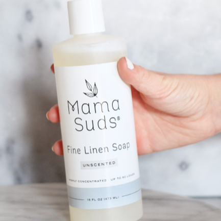 A hand holds a bottle of MamaSuds Fine Linen Soap, an unscented, highly concentrated, biodegradable laundry detergent, against a light gray background.
