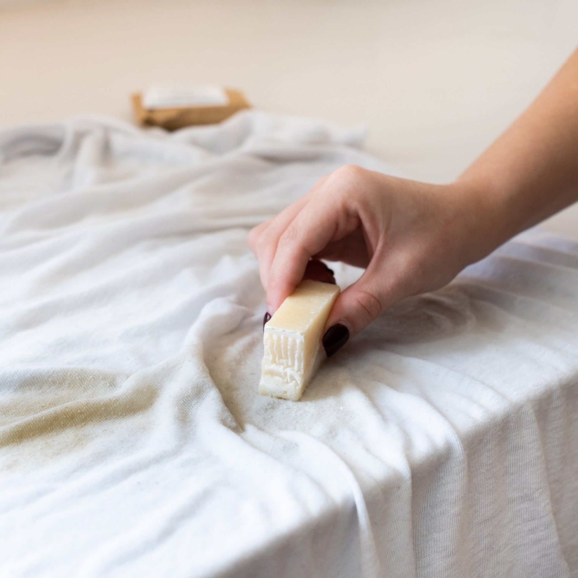 A hand uses the MamaSuds Laundry Stain Stick, a vegan and biodegradable bar, to clean a stain on white fabric. In the blurred background, another brown rectangular Laundry Stain Stick is visible.