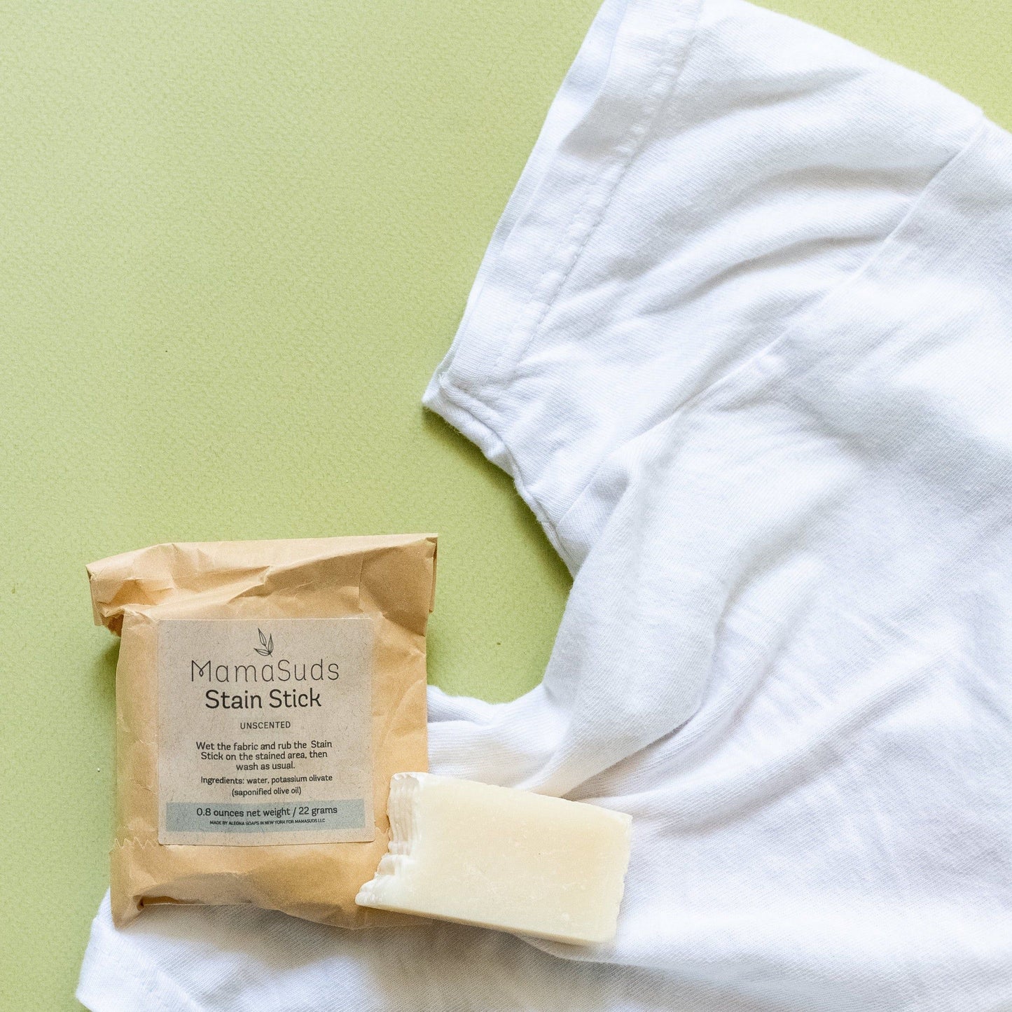A white T-shirt lies on a light green surface next to a brown package labeled MamaSuds Laundry Stain Stick and an unwrapped, vegan stain remover bar.