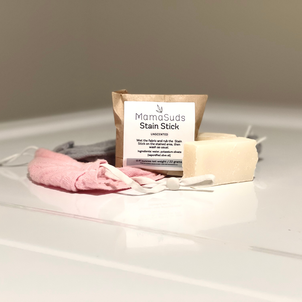 A MamaSuds Laundry Stain Stick, labeled in brown paper, sits on a white surface next to pink and gray reusable cloths, emphasizing this biodegradable laundry essential in a clean, minimalistic scene.