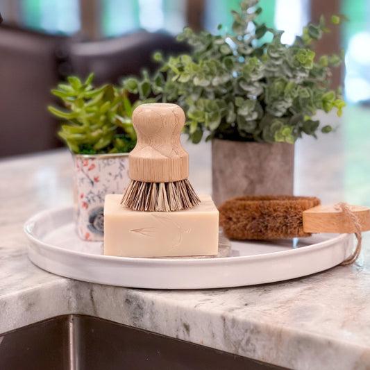 On a marble countertop, a round tray displays MamaSuds Solid Dish Soap (vegan and biodegradable), a wooden scrub brush, a brown cleaning brush, and two small potted plants. The softly blurred background adds an elegant touch.