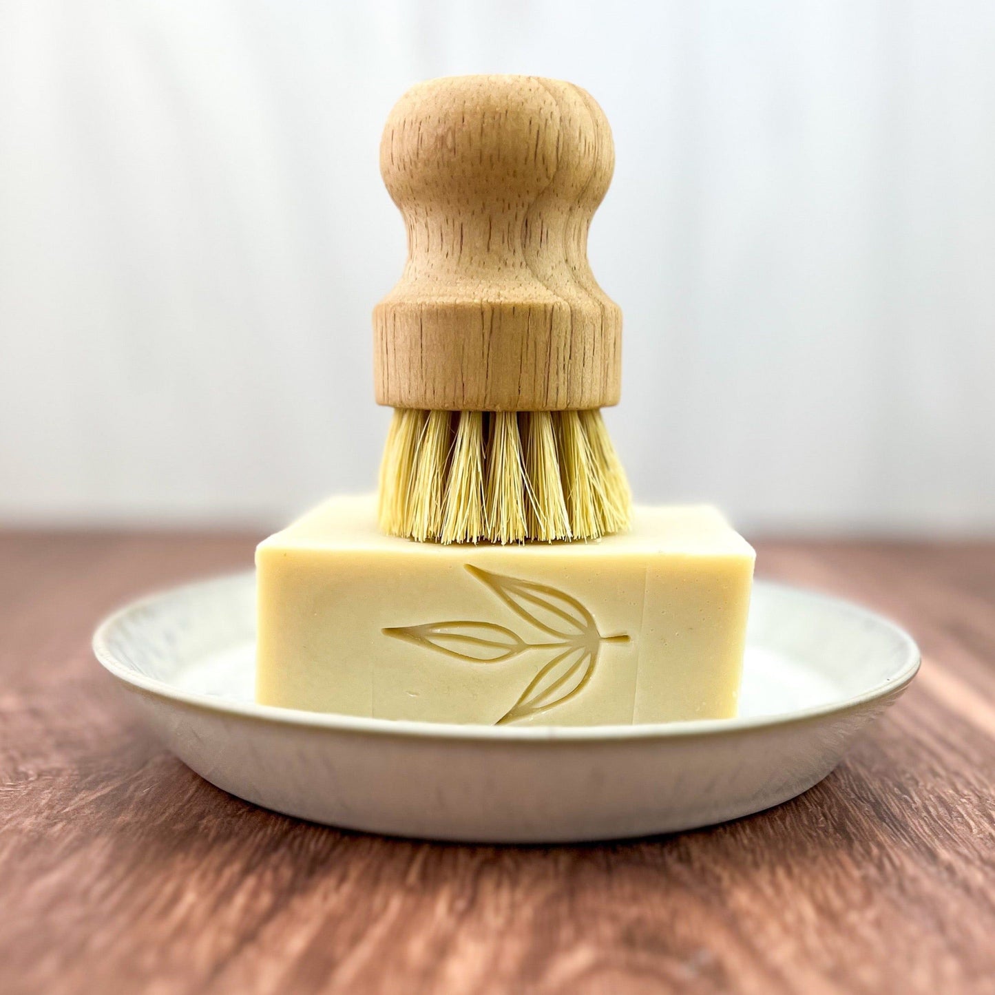 A wooden dish brush stands upright on a rectangular bar of MamaSuds Solid Dish Soap featuring a leaf design, all arranged on a white plate atop a wooden surface.