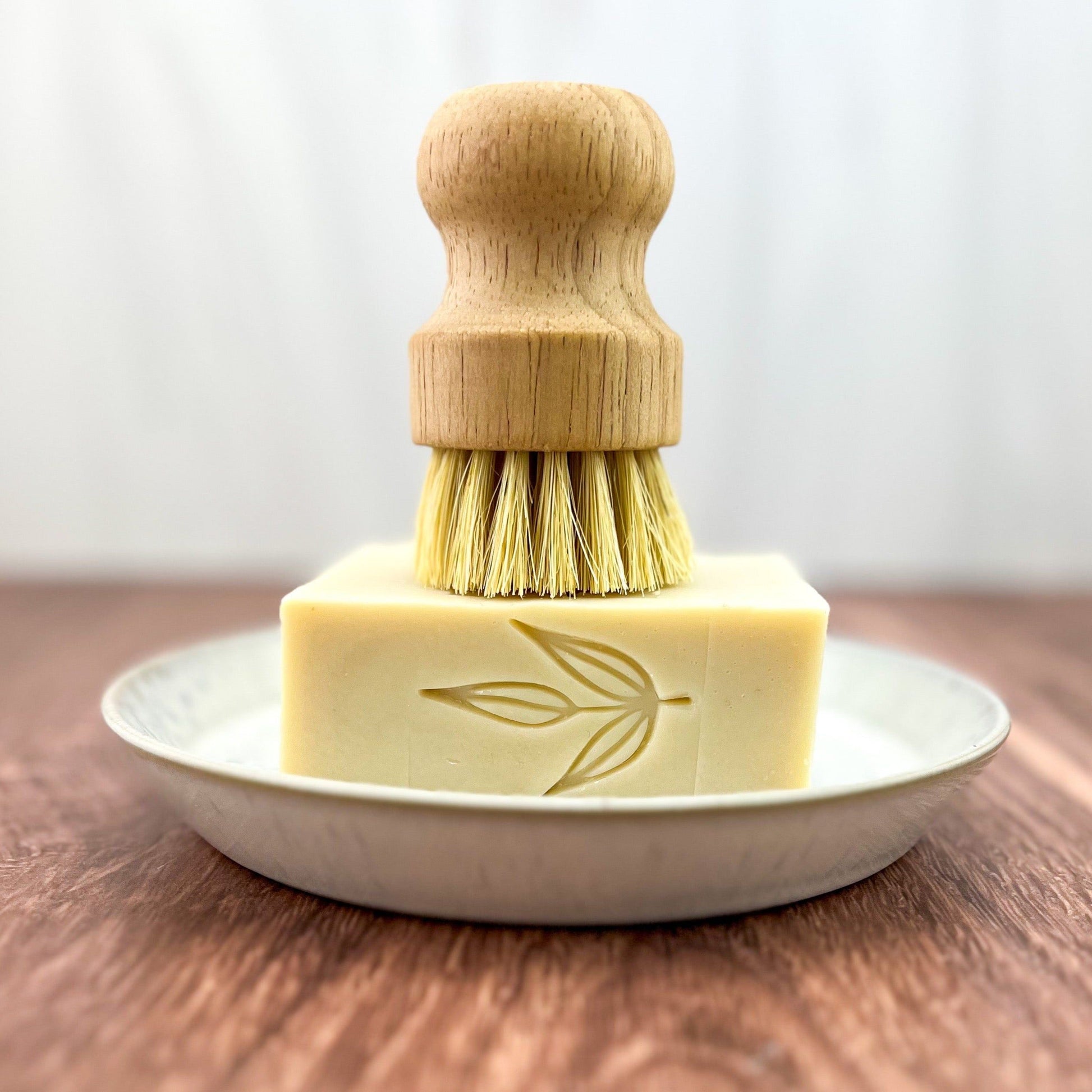 A wooden dish brush stands upright on a rectangular bar of MamaSuds Solid Dish Soap featuring a leaf design, all arranged on a white plate atop a wooden surface.