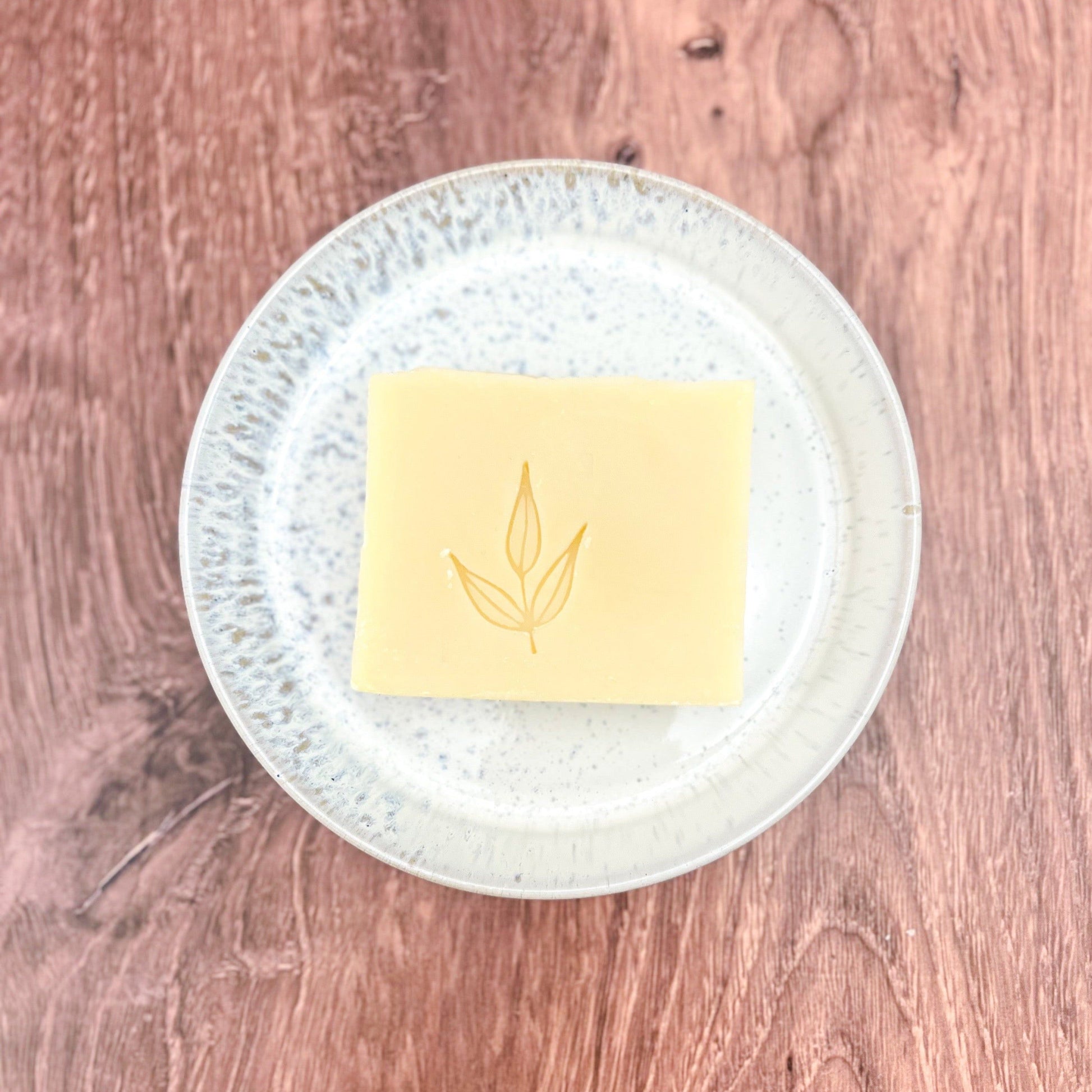 A square bar of MamaSuds Solid Dish Soap with a simple leaf design rests on a white speckled plate atop a wooden surface.