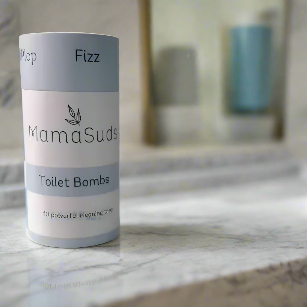 A cylindrical container labeled MamaSuds Toilet Bomb Cleaning Tabs | Non-toxic Toilet Cleaner sits on a marble bathroom counter, with blurred bathroom items in the background.