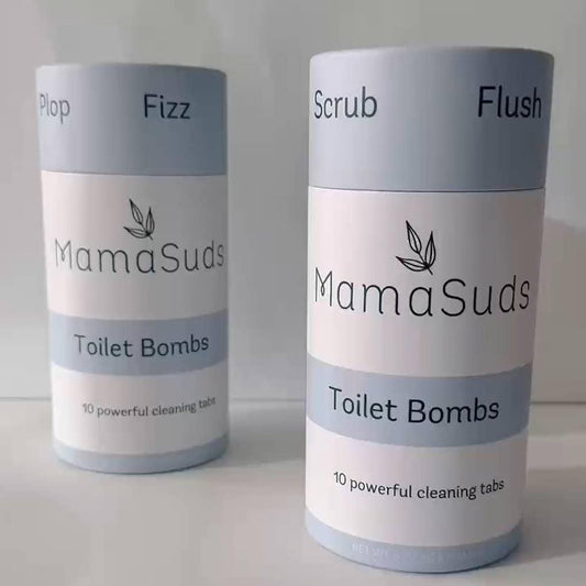 Two cylindrical MamaSuds Toilet Bomb Cleaning Tabs containers, in minimalist blue and white, stand side by side. Each holds 10 non-toxic, citric acid cleaning tabs—one labeled Plop Fizz, the other Scrub Flush.