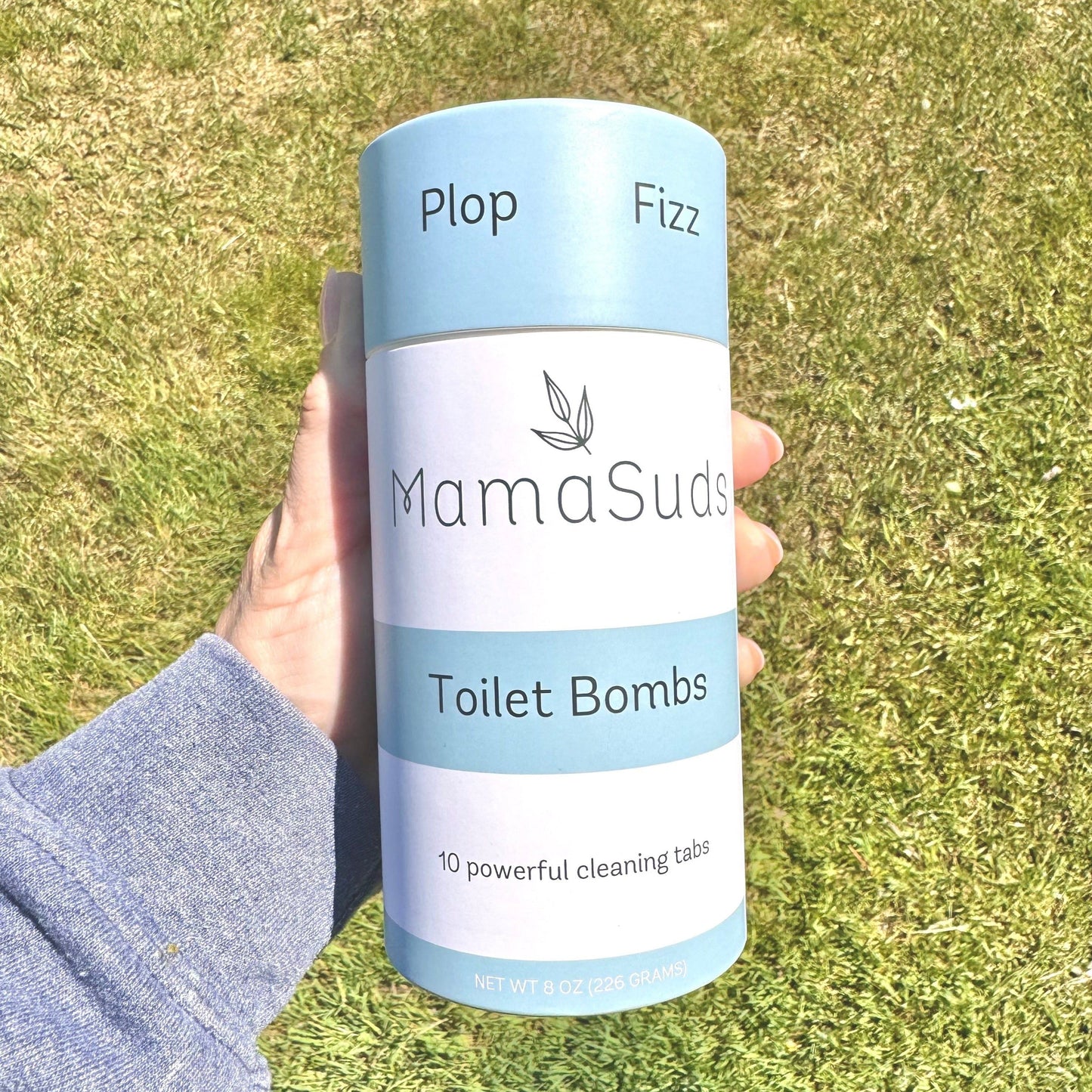 A hand holds a MamaSuds Toilet Bomb Cleaning Tabs | Non-toxic Toilet Cleaner pack with Plop and Fizz on top. The package shows 10 non-toxic cleaning tabs for toilet use, set outdoors on grass.