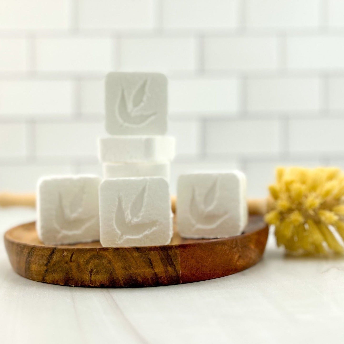 Five white, leaf-imprinted Toilet Bomb Cleaning Tabs from MamaSuds are stacked on a wooden dish in front of a soft-focus brush and white subway tiles—ideal for non-toxic toilet cleaning.
