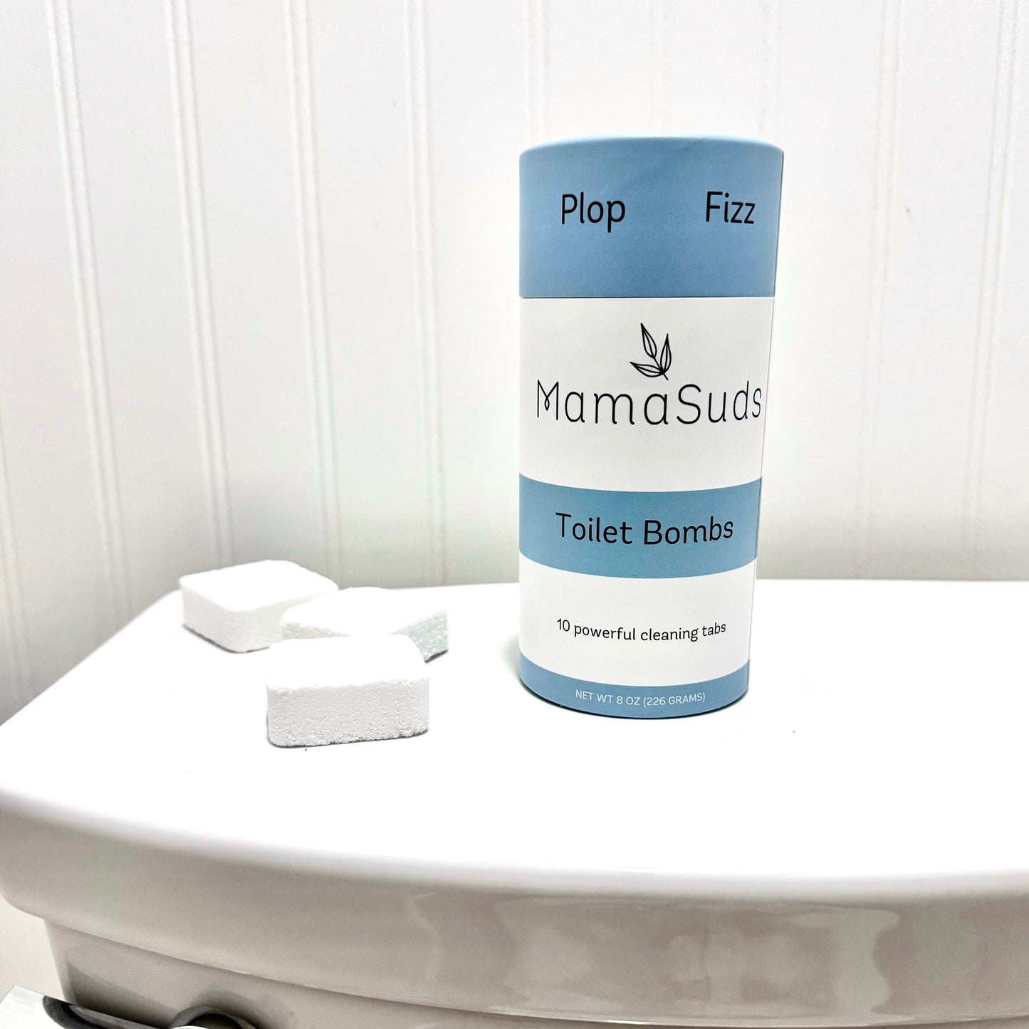 A cylindrical container labeled MamaSuds Toilet Bomb Cleaning Tabs | Non-toxic Toilet Cleaner sits on a toilet lid, with three white tabs nearby. The blue and white packaging highlights “10 powerful cleaning tabs” for non-toxic cleaning.