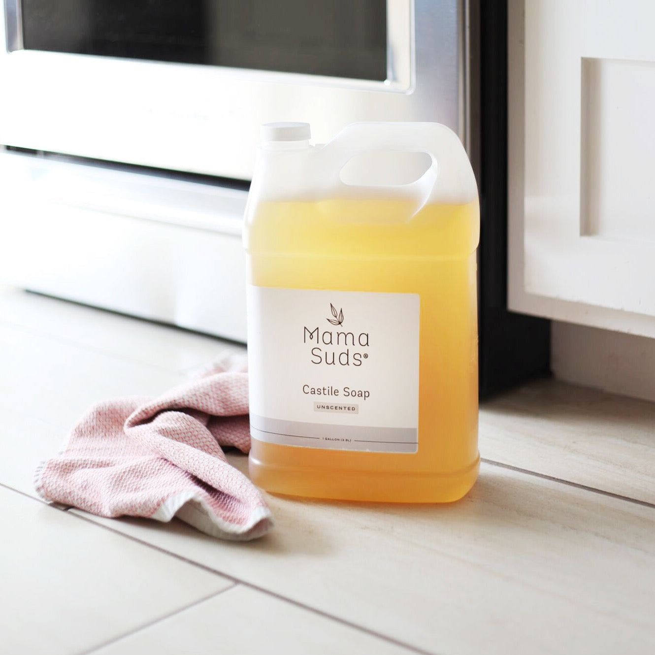 A large jug of MamaSuds Castile Liquid Soap, an all-purpose cleaner made with pure essential oils, sits on a light tiled floor next to a pink cloth in front of an oven and white cabinet.