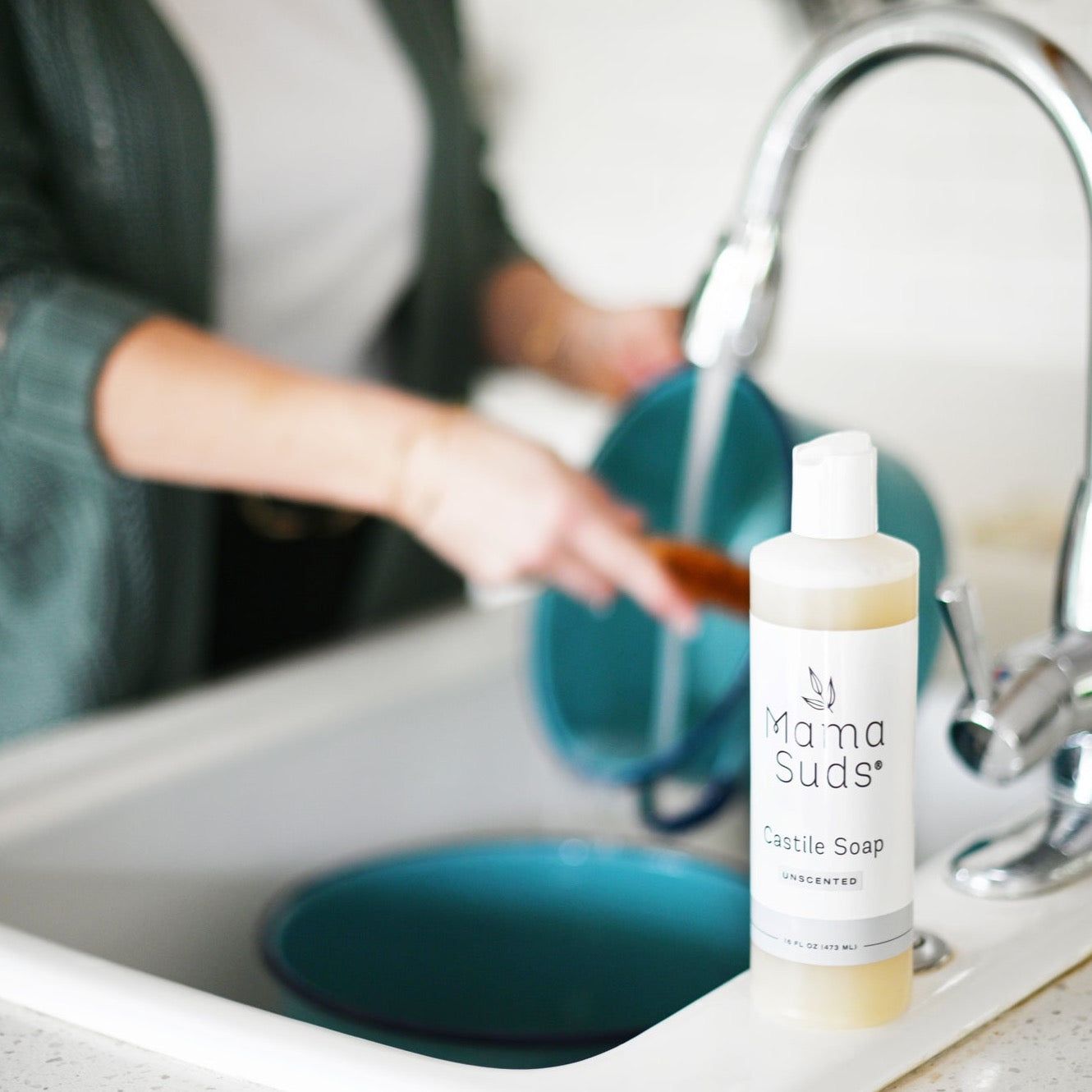 A bottle of MamaSuds Castile Liquid Soap, crafted with pure essential oils, sits on a kitchen sink as someone washes blue pans in the background, showcasing this all-purpose cleaner as the highlight of the kitchen.