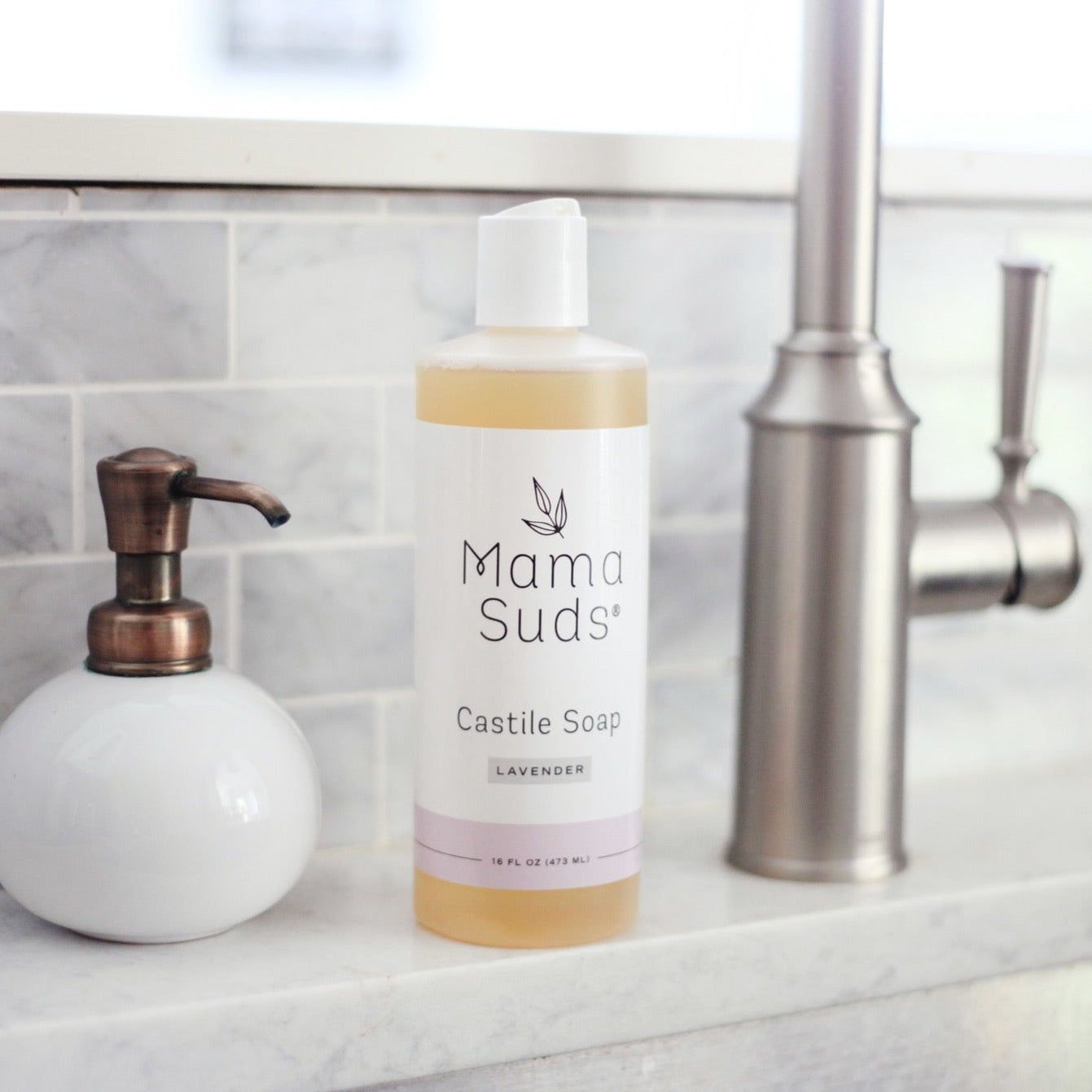 A bottle of MamaSuds Castile Liquid Soap made with pure essential oils sits on a marble countertop beside a white soap dispenser and silver faucet in a modern kitchen or bathroom.