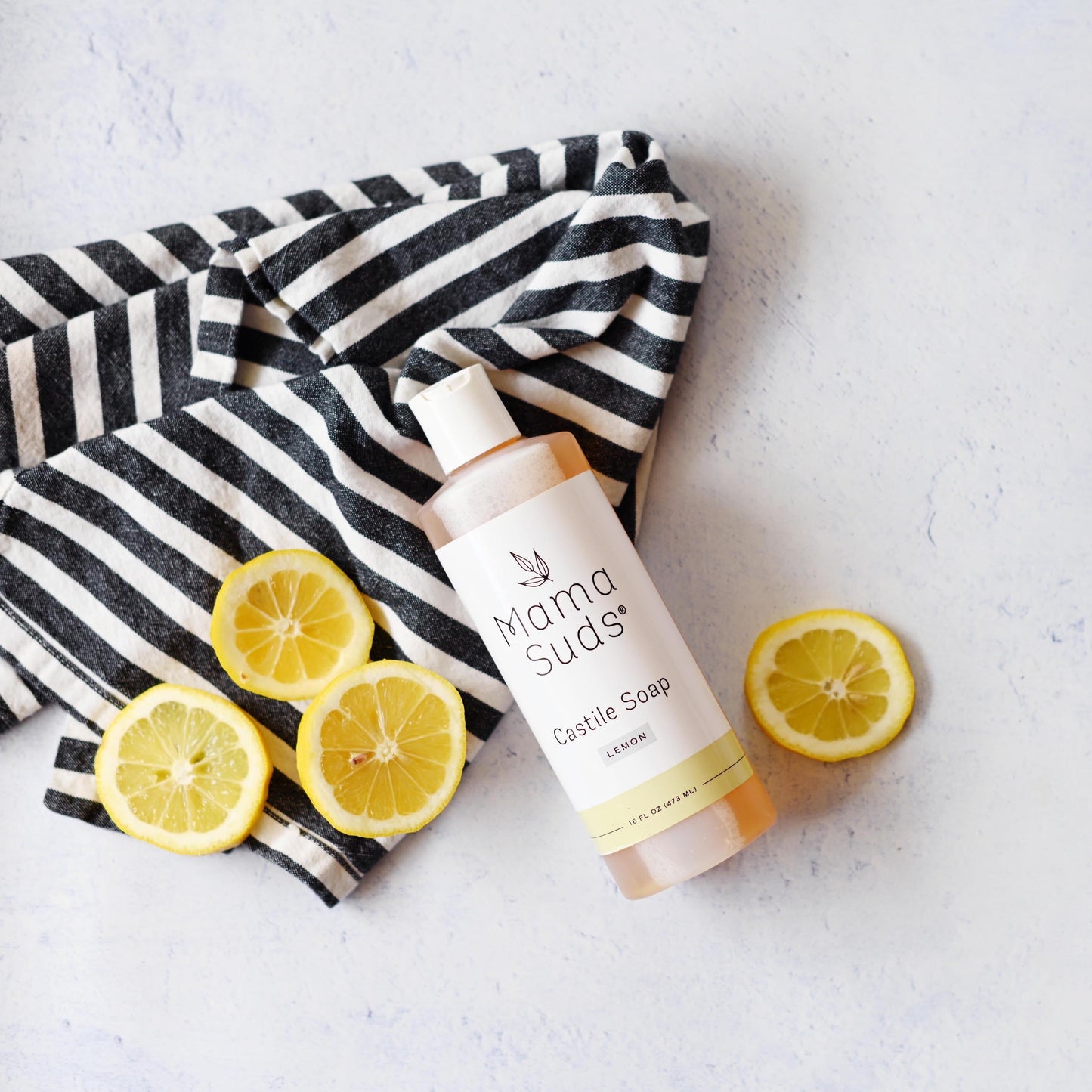 A bottle of MamaSuds Castile Liquid Soap labeled Lemon rests on a black and white striped cloth, surrounded by sliced lemons and pure essential oils, highlighting this all-purpose cleaner on a light-colored surface.