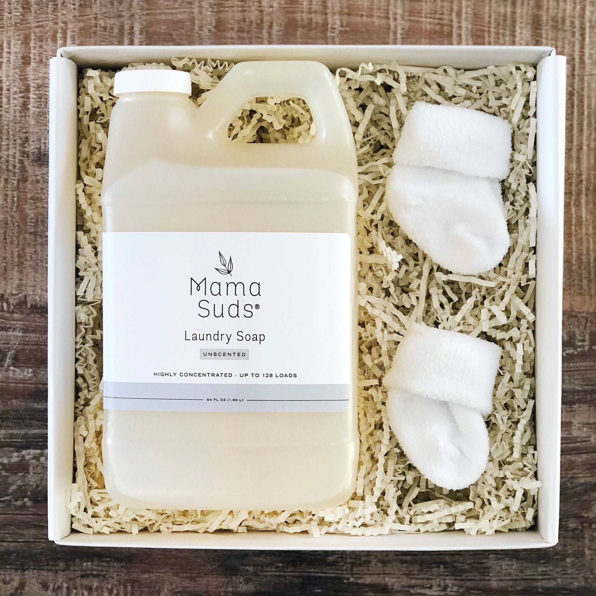 A box with a large bottle of MamaSuds Laundry Detergent Soap and a pair of small white baby socks, placed on shredded paper filler.