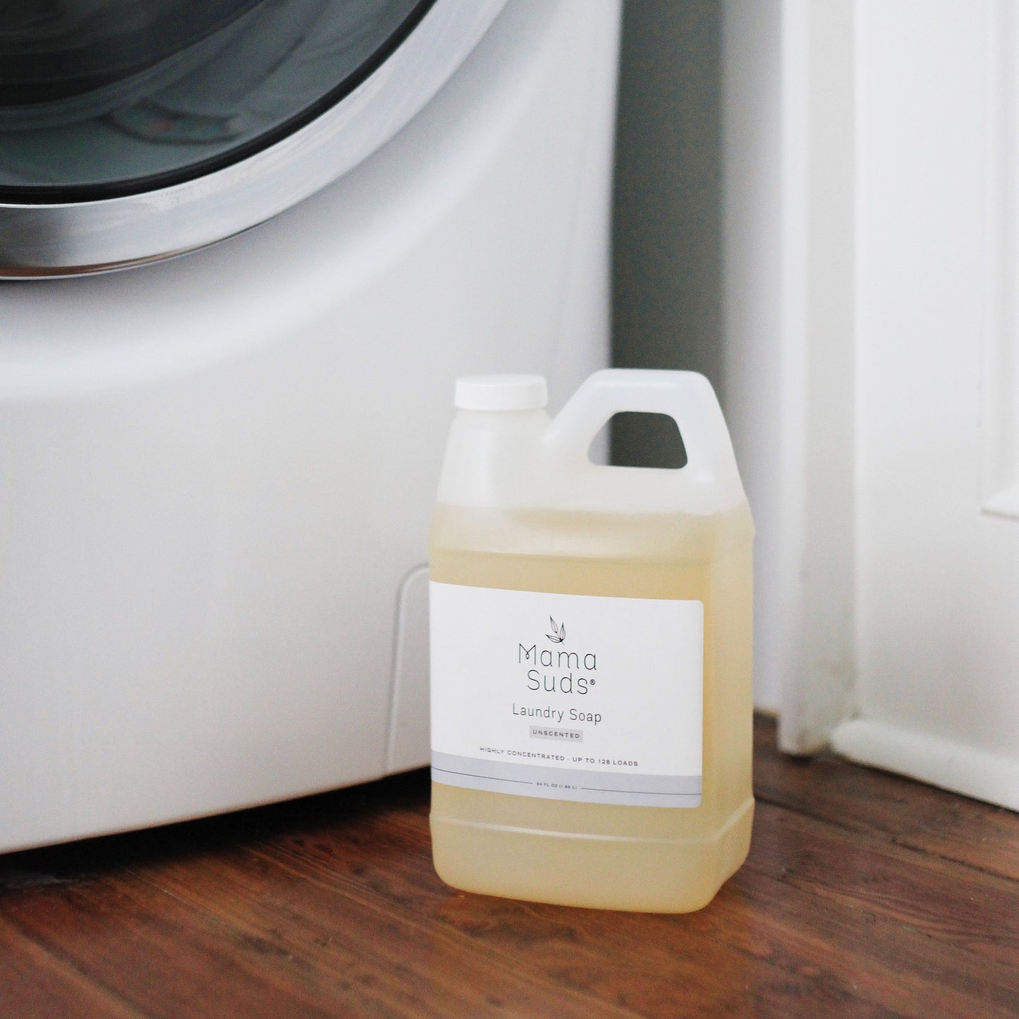 A container of MamaSuds Laundry Detergent Soap, made with natural ingredients, sits on a wooden floor next to a white washing machine.
