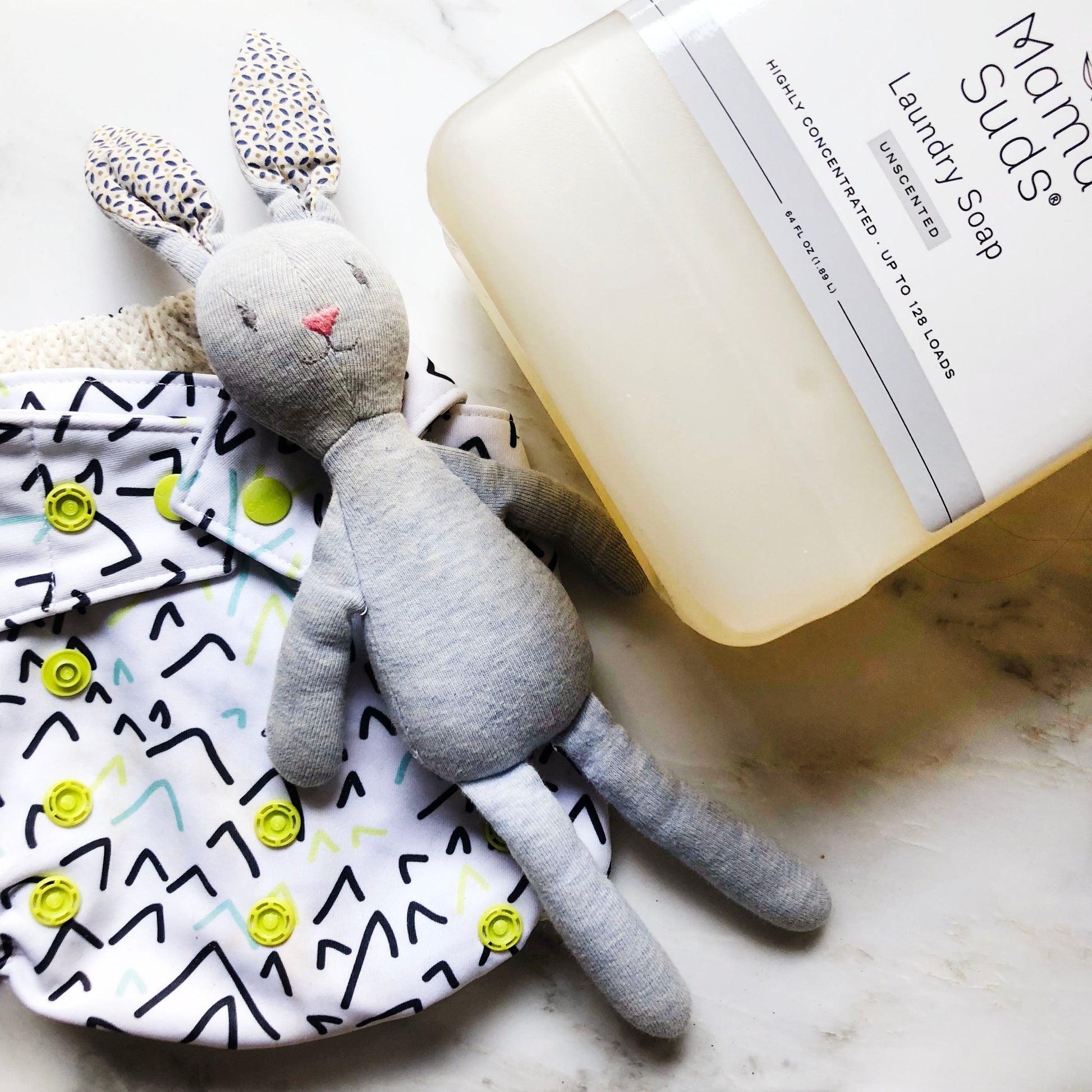 A gray stuffed rabbit, a patterned cloth diaper with yellow snaps, and a large bottle of MamaSuds Laundry Detergent Soap are arranged on a white surface.