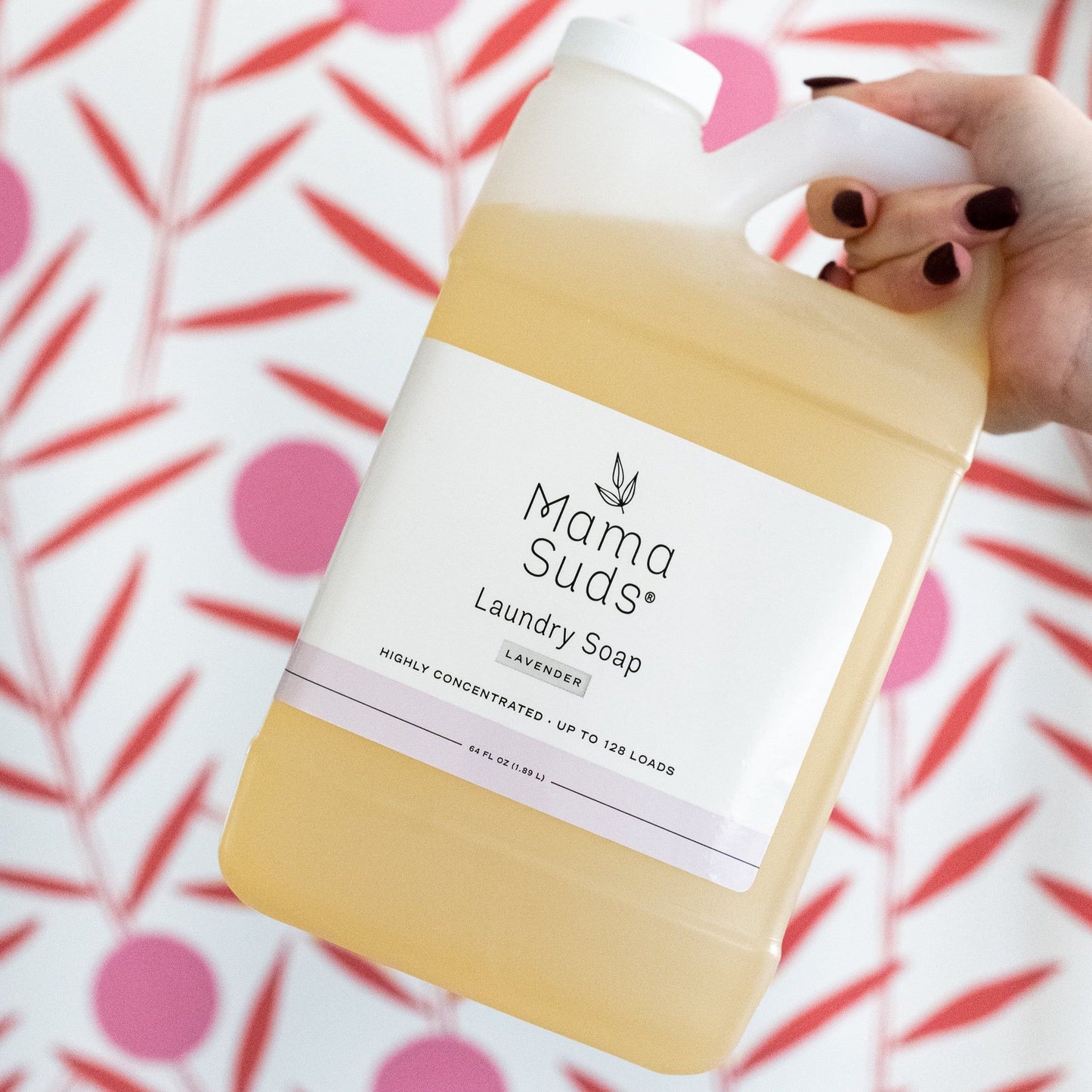 A hand with dark-painted nails holds a large bottle of MamaSuds Laundry Detergent Soap in front of a pink and red patterned background. The label highlights its eco-friendly, nontoxic formula and high concentration for up to 128 loads.