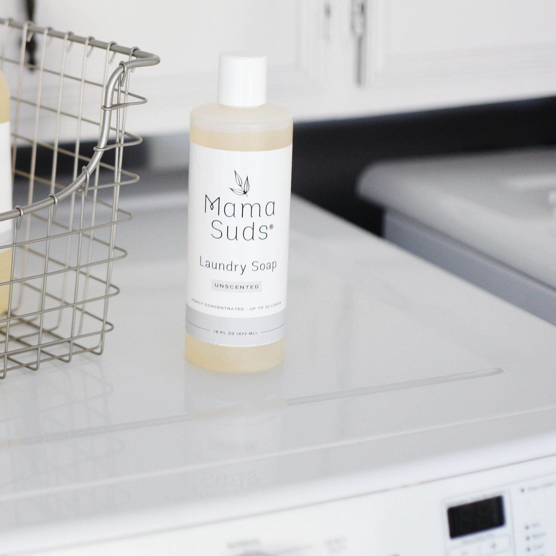 A bottle of MamaSuds Laundry Detergent Soap, an eco-friendly and nontoxic cleaner, sits atop a white washing machine beside a wire basket in the laundry room.