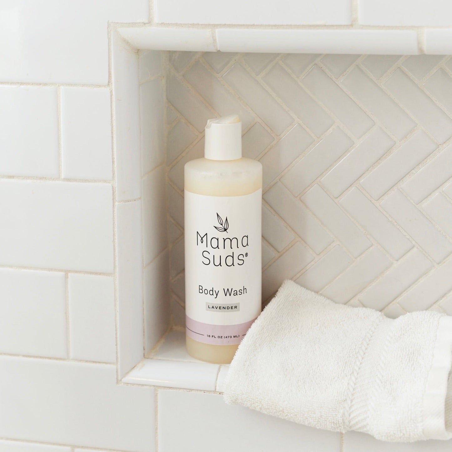 A MamaSuds Body Wash Soap bottle rests on a white tiled shower shelf beside a folded white towel, with a herringbone tile pattern in the background.