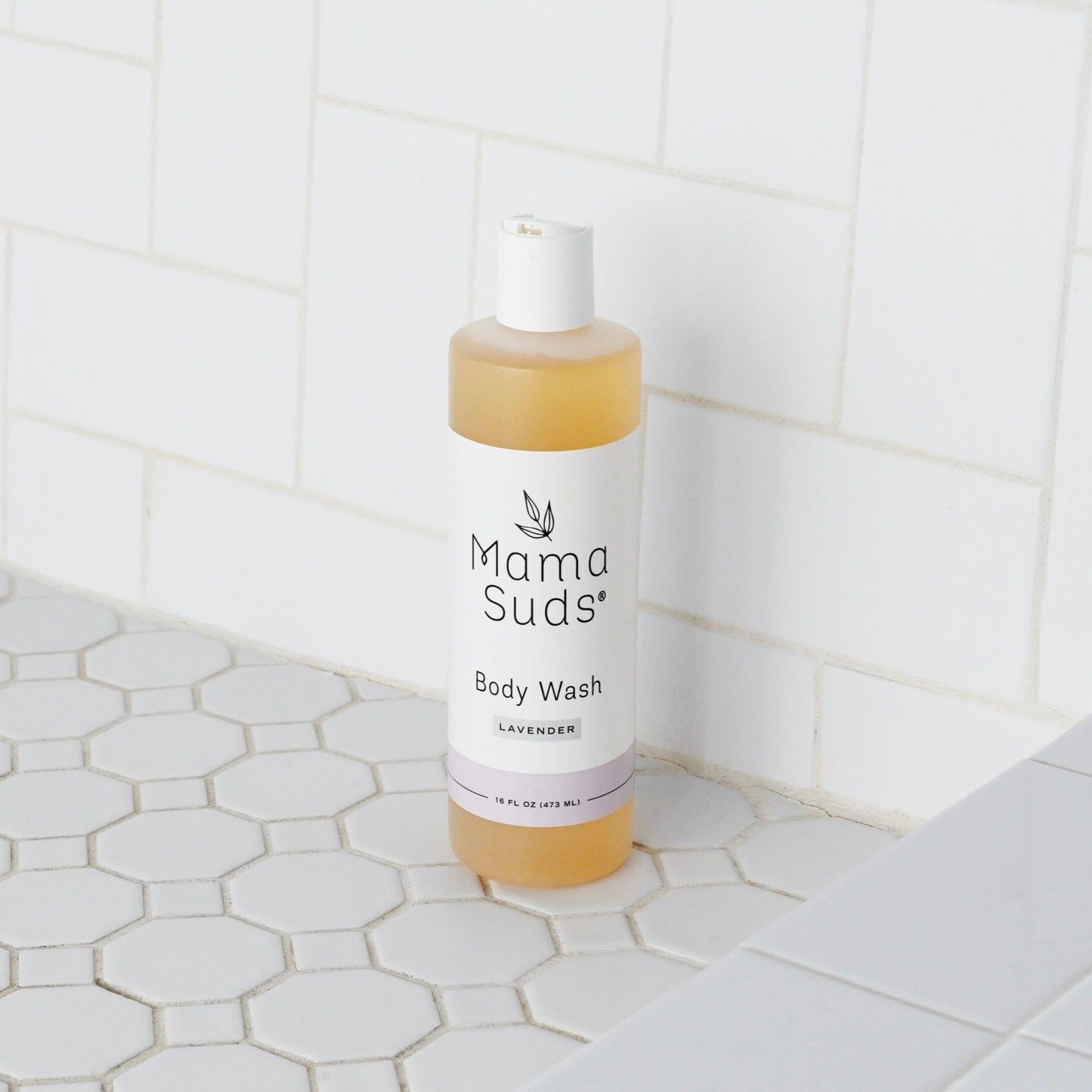A bottle of MamaSuds Body Wash Soap, made with pure castile soap and essential oils, sits on a white hexagonal-tiled shower floor against white rectangular tiles. The label features black text and a small leaf graphic.