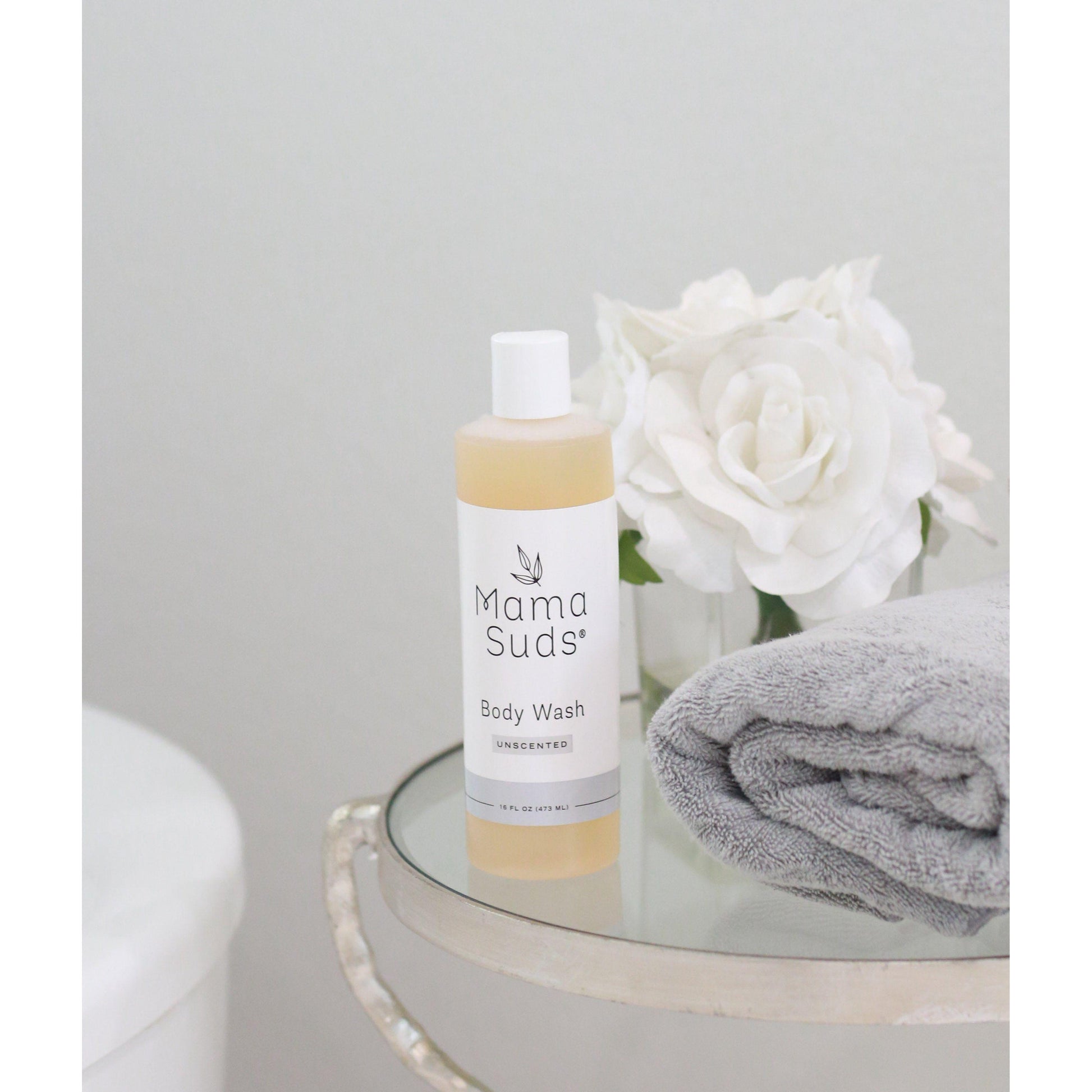 A bottle of MamaSuds Body Wash Soap, made from saponified olive oil castile soap, rests on a glass table next to a folded gray towel and white roses, set against a minimal light background.