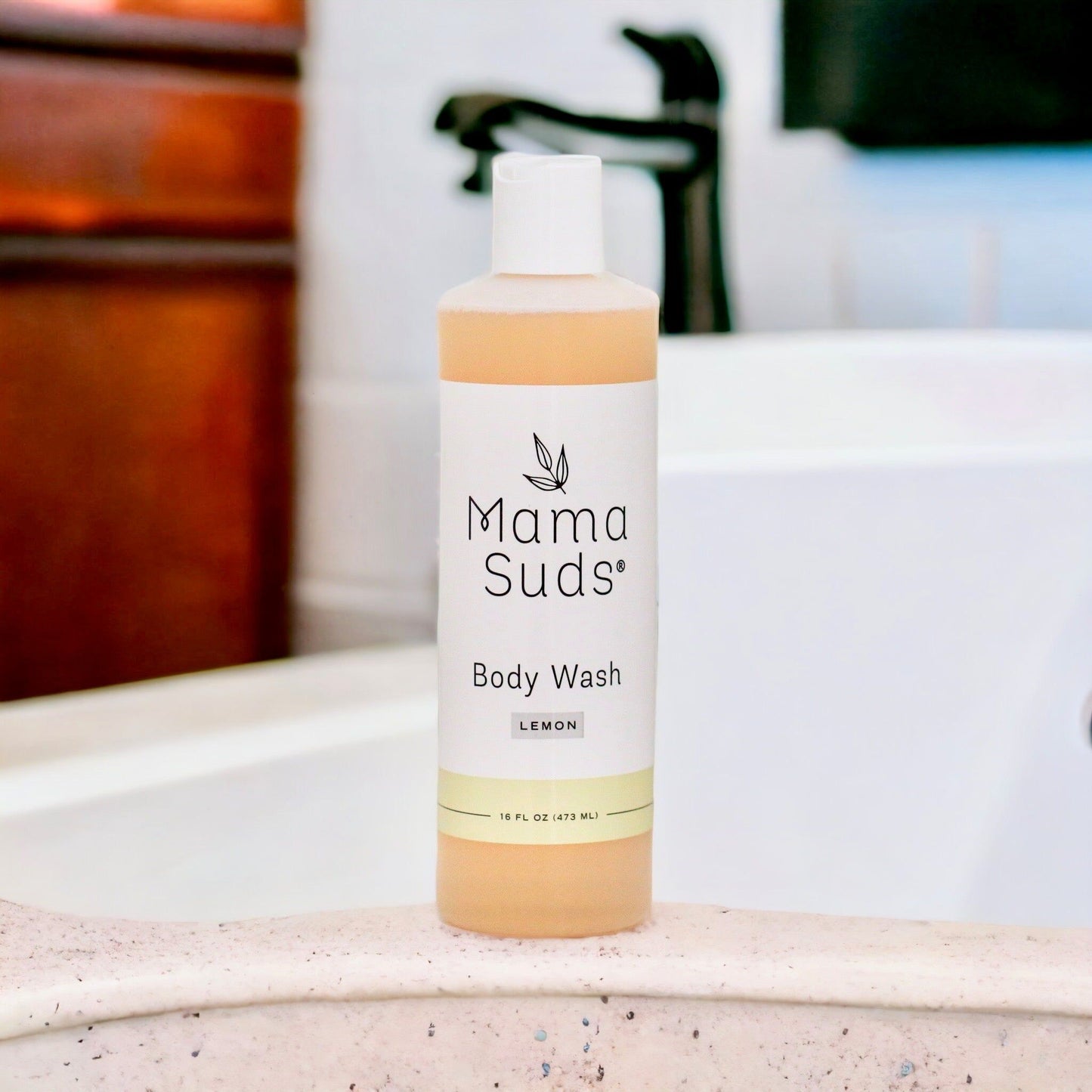 A bottle of MamaSuds Body Wash Soap rests on the edge of a bathtub, with a blurred faucet and bathroom fixtures in the background.