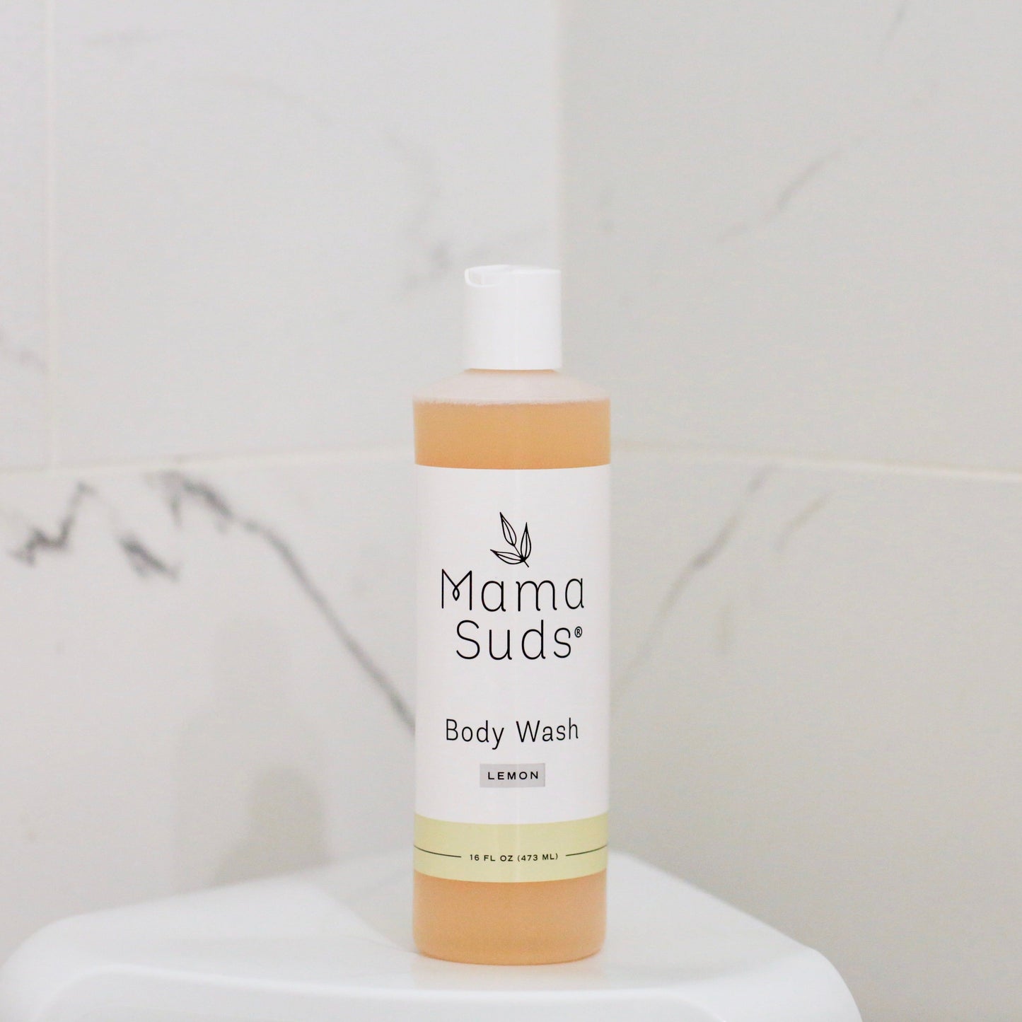 A bottle of MamaSuds Body Wash Soap, made with pure castile soap and saponified olive oil, sits on a white surface in front of a light marble-patterned tiled wall.