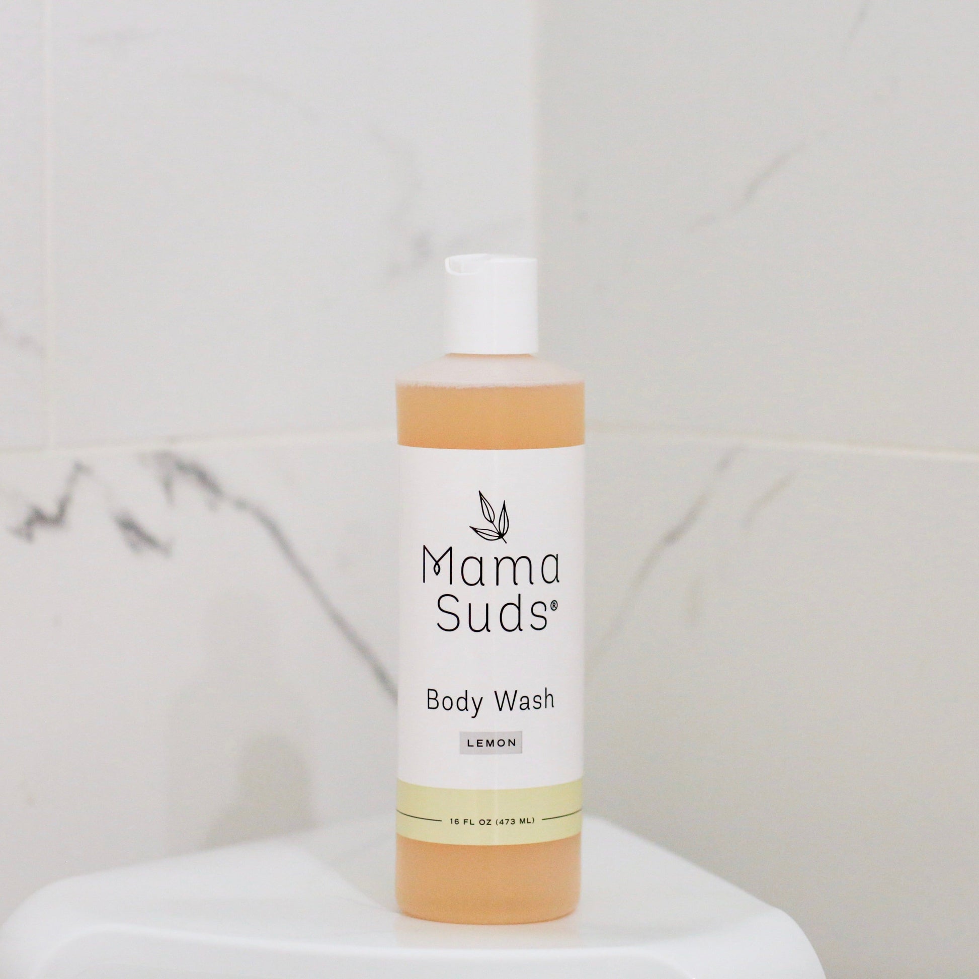 A bottle of MamaSuds Body Wash Soap, made with pure castile soap and saponified olive oil, sits on a white surface in front of a light marble-patterned tiled wall.