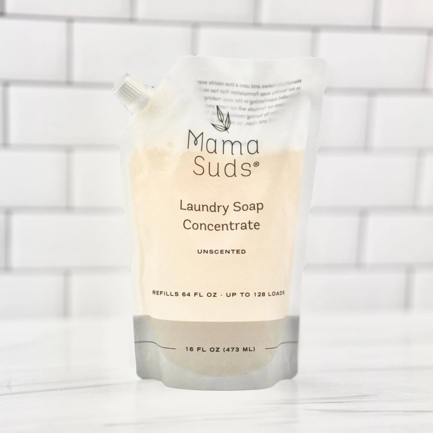 A recyclable MamaSuds Laundry Soap Concentrate Pouch, unscented and biodegradable, stands on a white surface with a tiled background. The 16 fl oz (473 ml) pouch is labeled for up to 128 loads.