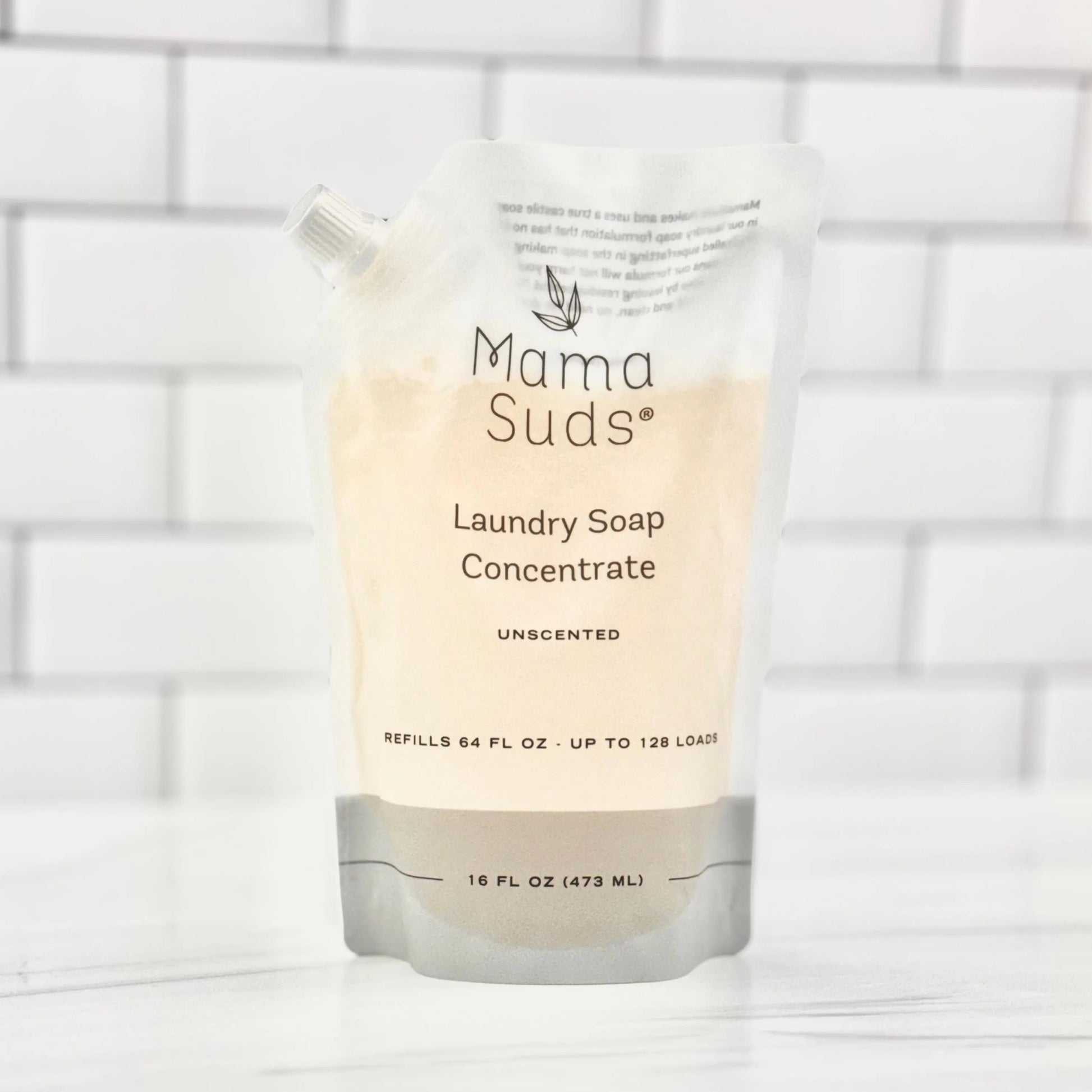 A recyclable MamaSuds Laundry Soap Concentrate Pouch, unscented and biodegradable, stands on a white surface with a tiled background. The 16 fl oz (473 ml) pouch is labeled for up to 128 loads.