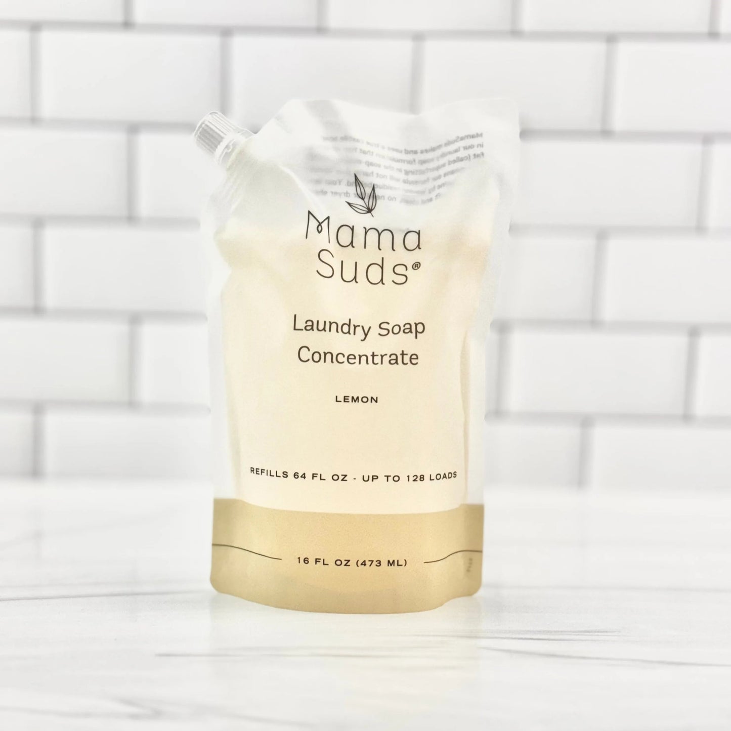 A white and beige biodegradable pouch of MamaSuds Laundry Soap Concentrate (Lemon), 16 fl oz, sits on a white surface with a tiled background; the recyclable pouch refills up to 128 loads.