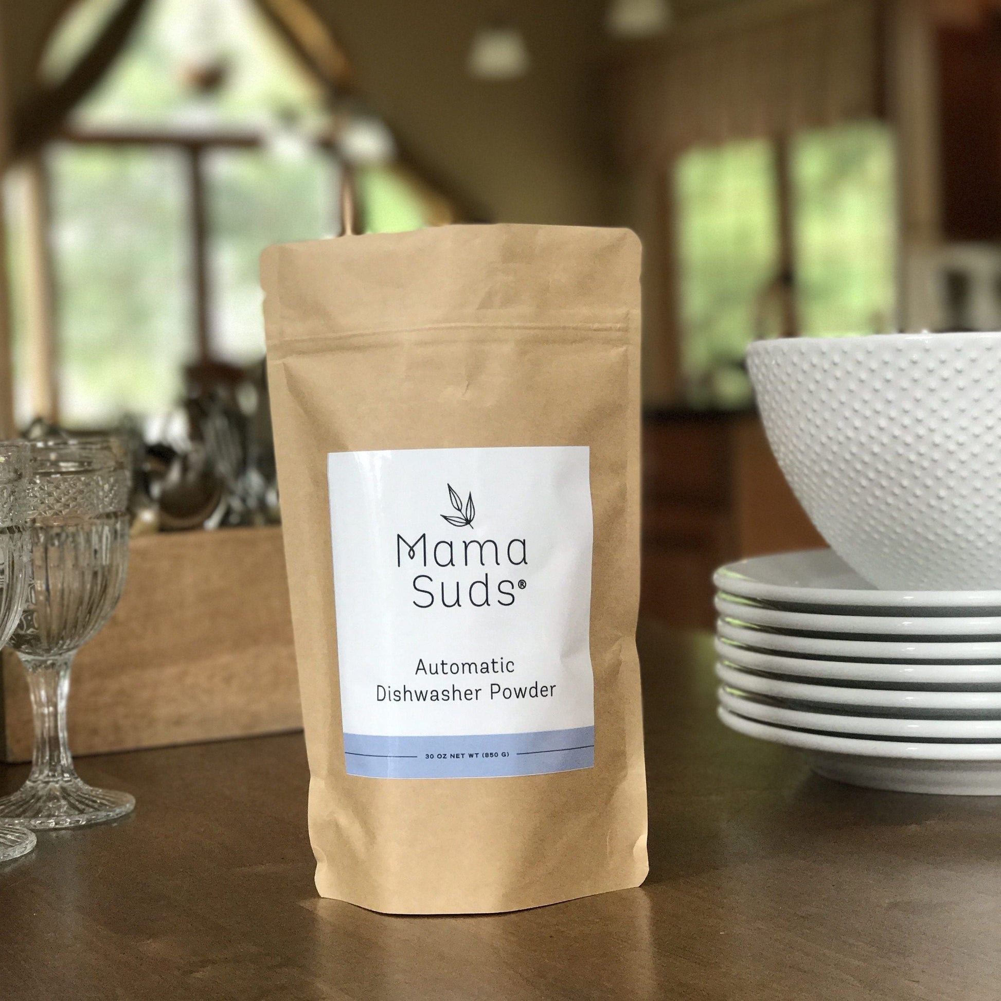 A brown paper bag labeled MamaSuds Automatic Dishwasher Powder | Nontoxic and Eco-Friendly sits on a wooden table beside stacked white bowls, glassware, and a blurred kitchen background.