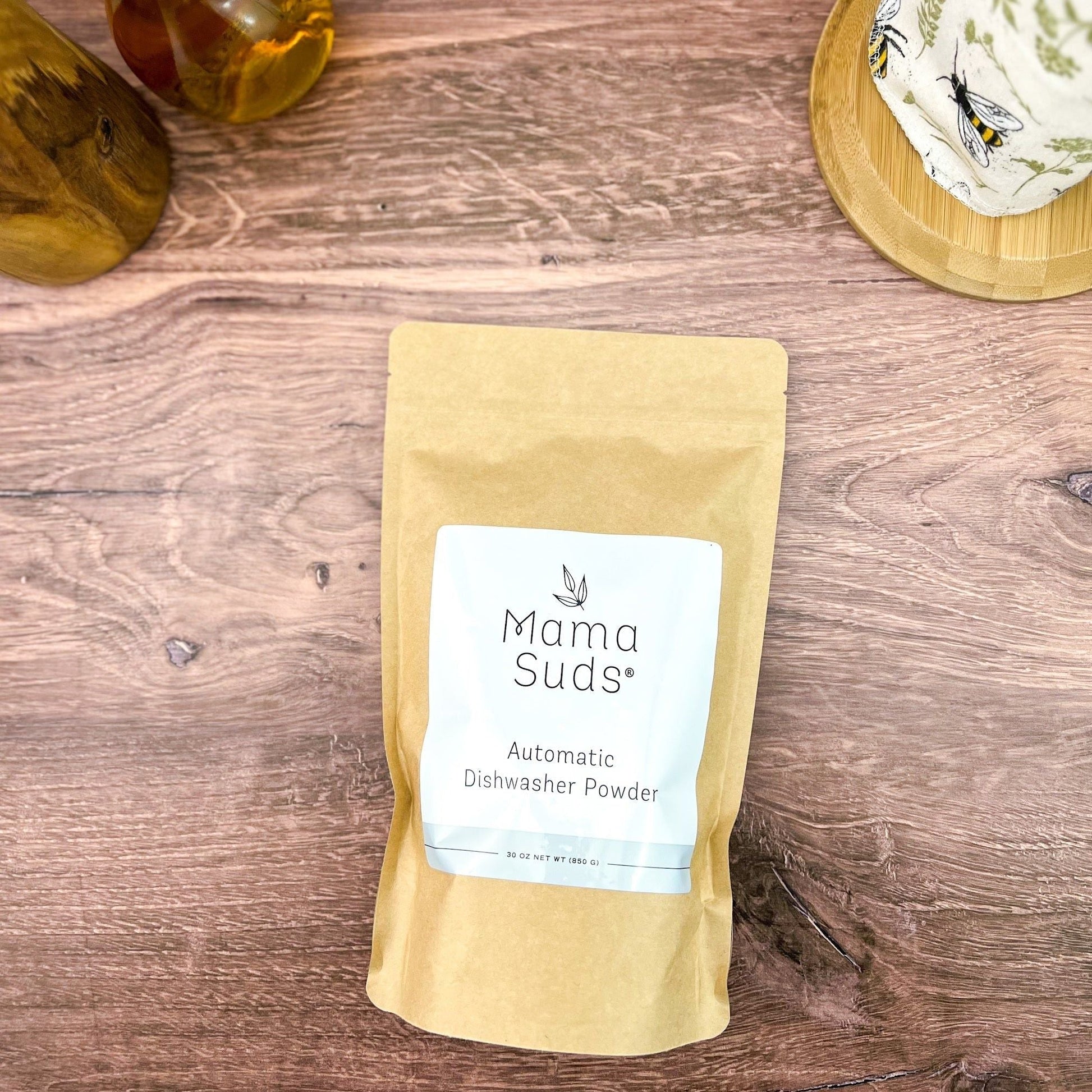 A brown paper pouch labeled MamaSuds Automatic Dishwasher Powder | Nontoxic and Eco-Friendly sits on a wooden surface among glass bottles and a covered container, offering a biodegradable, sustainable cleaning solution.