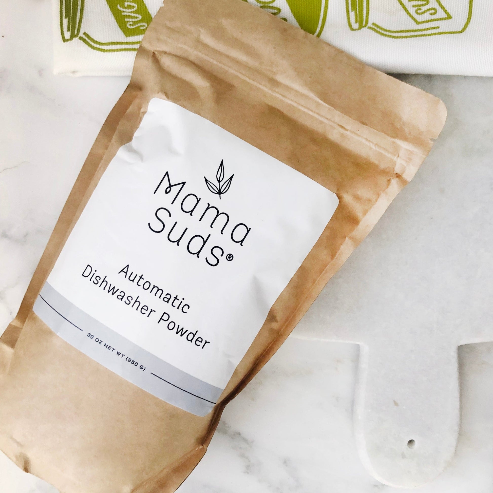 A brown paper pouch labeled MamaSuds Automatic Dishwasher Powder | Nontoxic and Eco-Friendly, an eco-friendly, biodegradable dishwasher powder, sits on a white surface with a green and white cloth in the background.