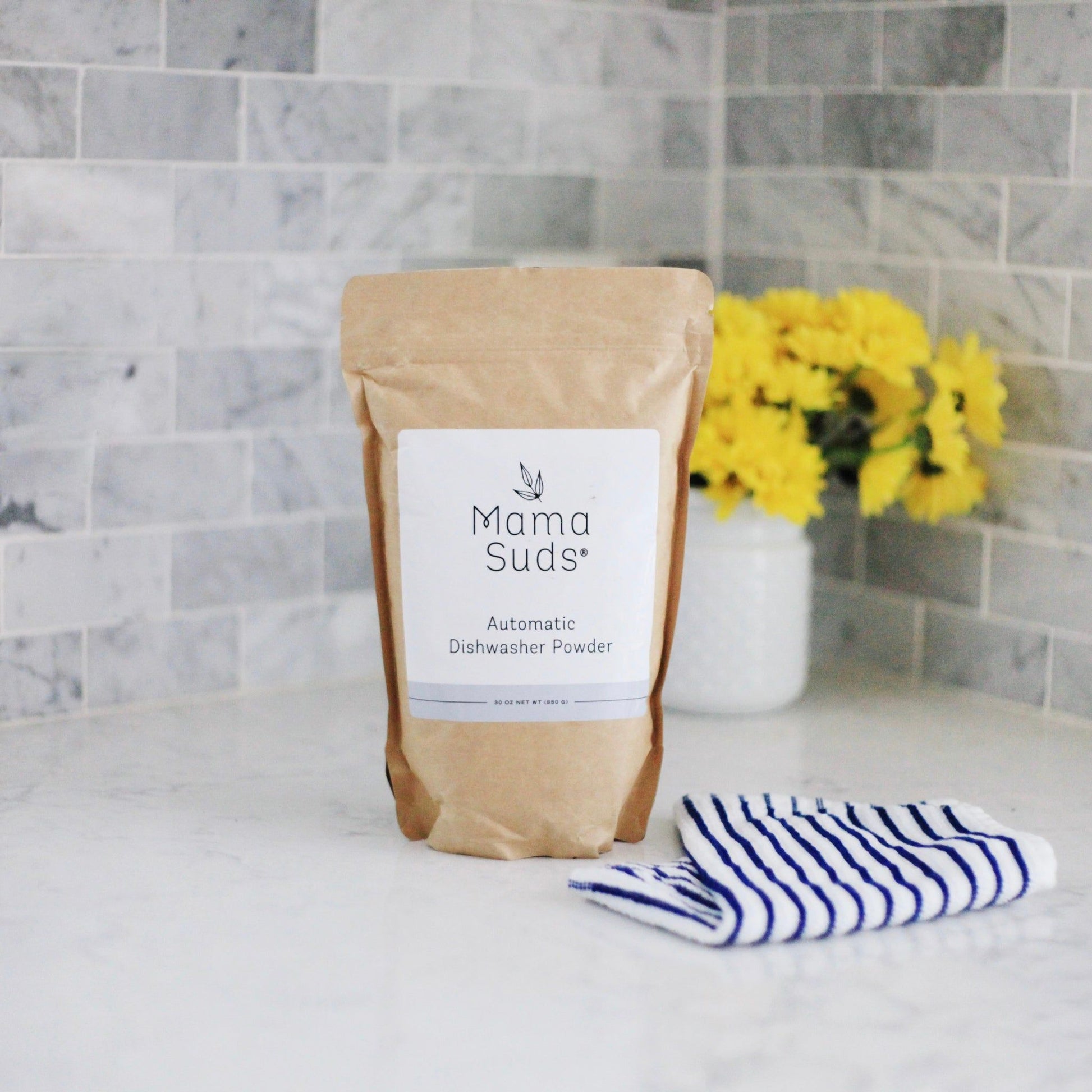 A brown paper bag labeled MamaSuds Automatic Dishwasher Powder | Nontoxic and Eco-Friendly sits on a marble countertop, next to yellow flowers in a white vase and a blue-striped towel.