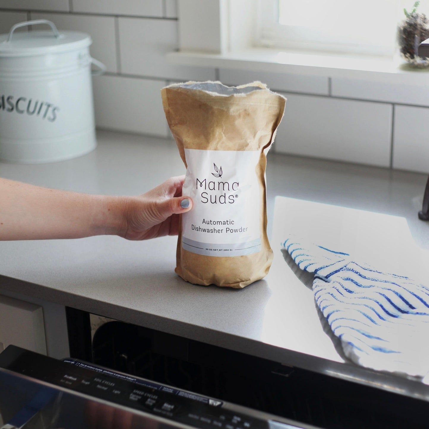 A hand holds a brown paper bag of MamaSuds Automatic Dishwasher Powder | Nontoxic and Eco-Friendly on a kitchen counter beside a dishwasher and a blue-striped dish towel.