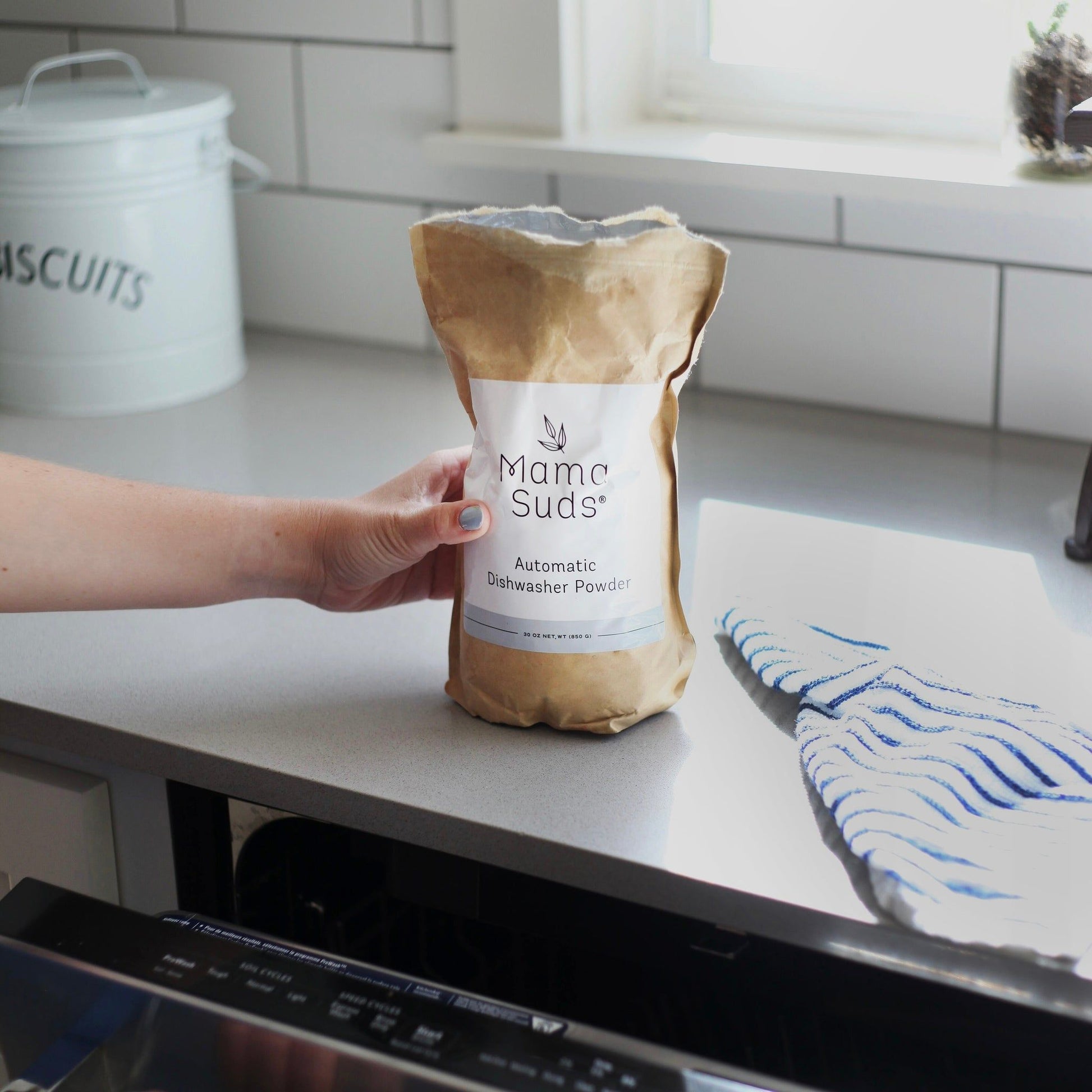 A hand holds a brown paper bag of MamaSuds Automatic Dishwasher Powder | Nontoxic and Eco-Friendly on a kitchen counter beside a dishwasher and a blue-striped dish towel.