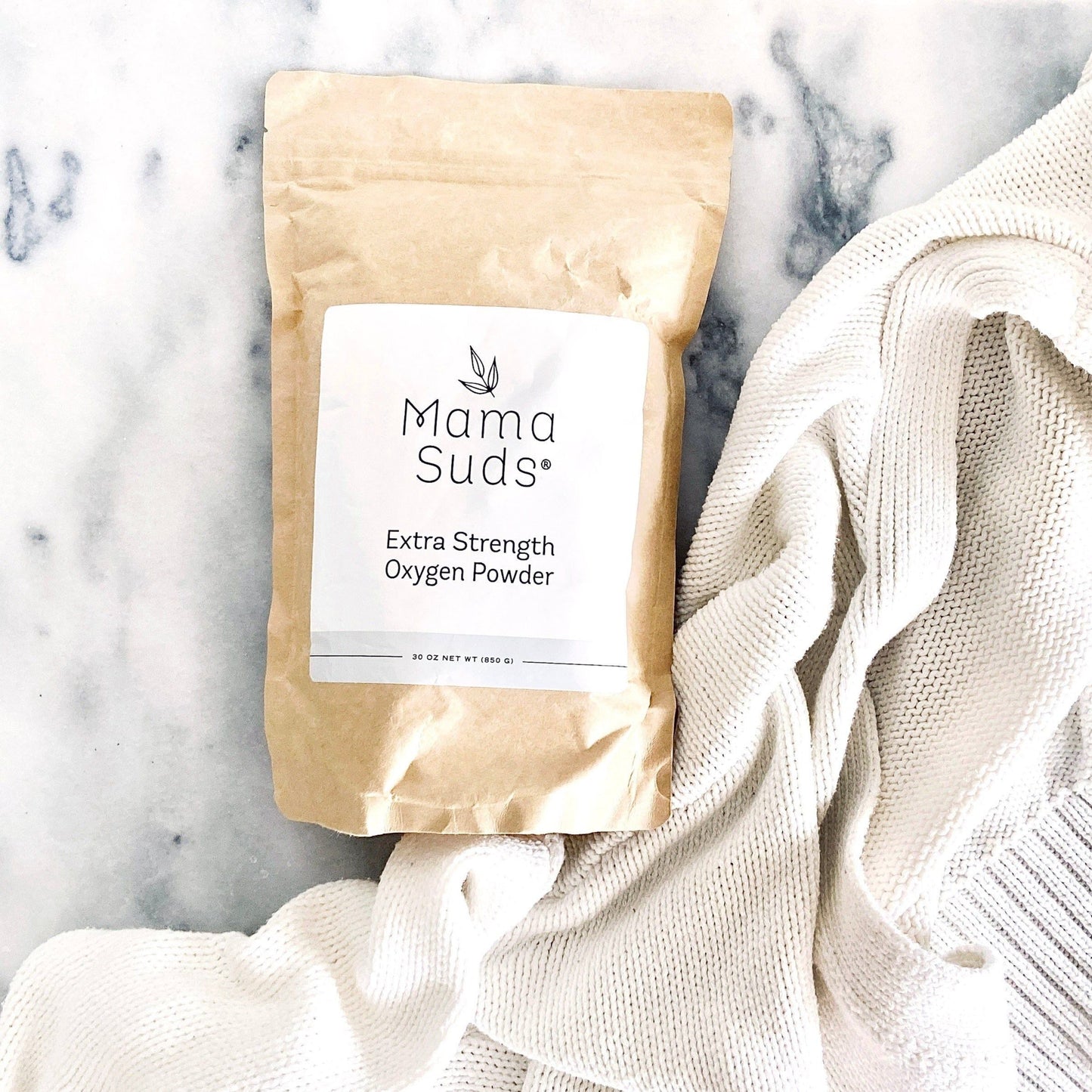 A brown paper bag labeled MamaSuds Extra Strength Oxygen Powder, a potent bleach alternative and stain remover, rests on a white marble surface beside a textured knit fabric.
