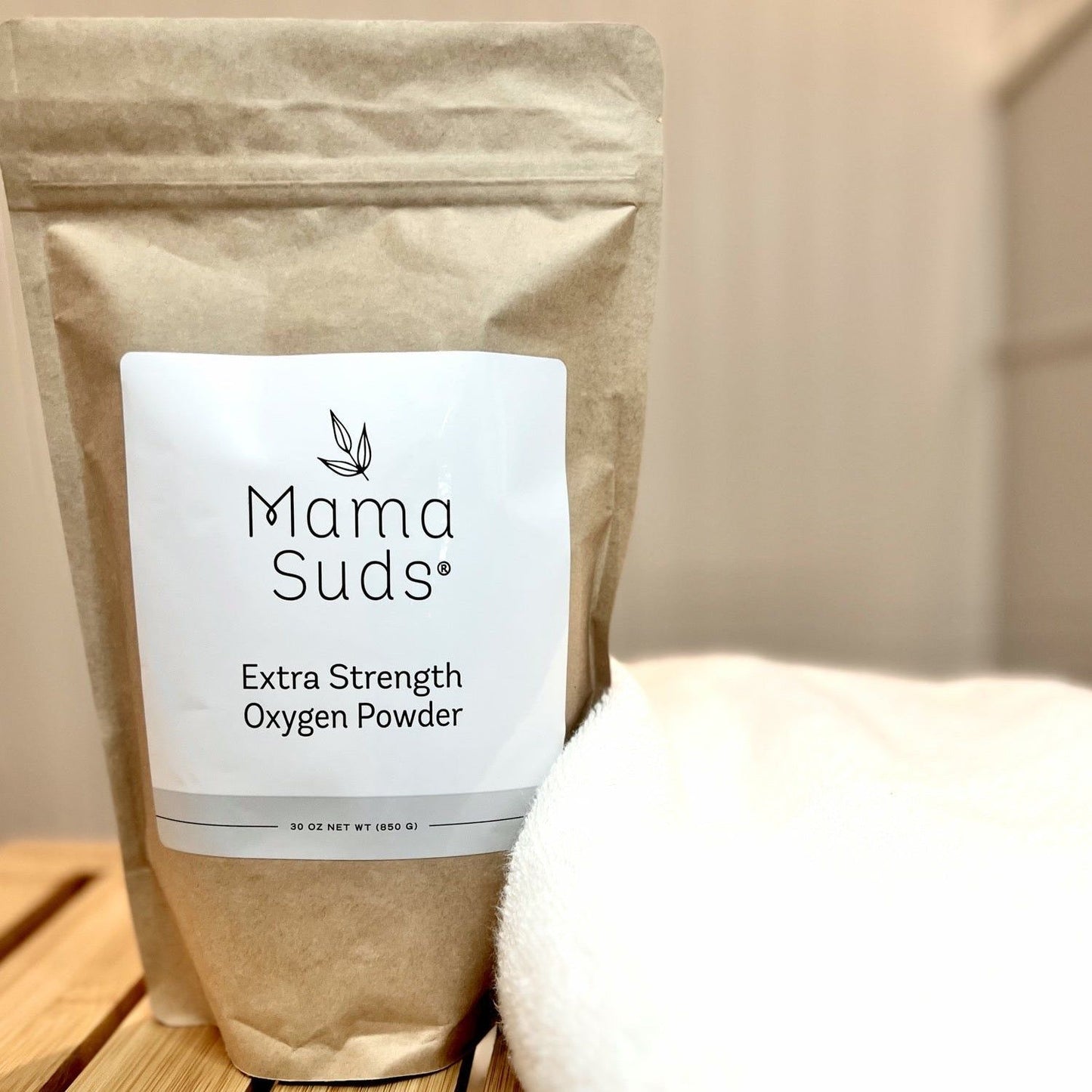 A brown paper bag labeled MamaSuds Extra Strength Oxygen Powder, a bleach alternative and stain remover, sits on a wooden surface beside a folded white towel with a softly blurred background.