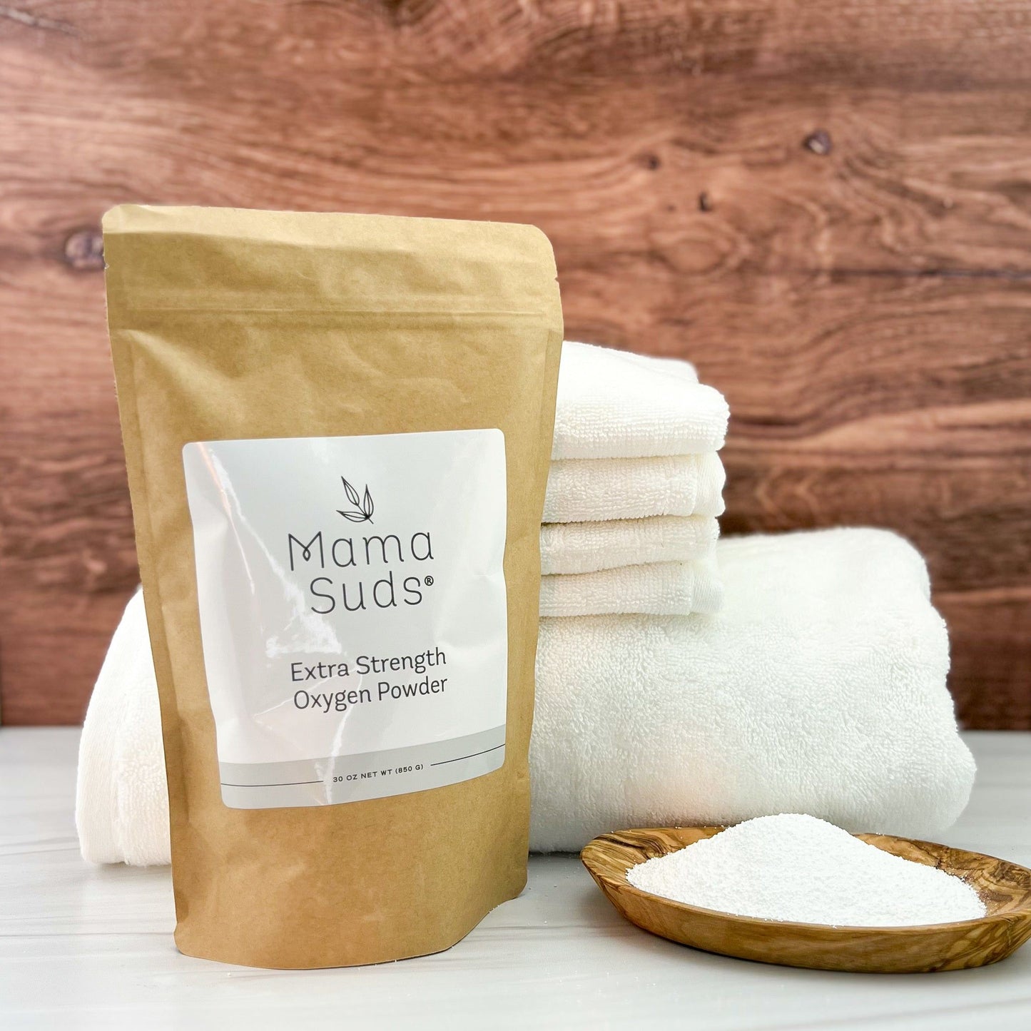 A brown bag labeled MamaSuds Extra Strength Oxygen Powder, a powerful bleach alternative and stain remover, stands beside a wooden dish of white powder and folded towels on a wooden background.