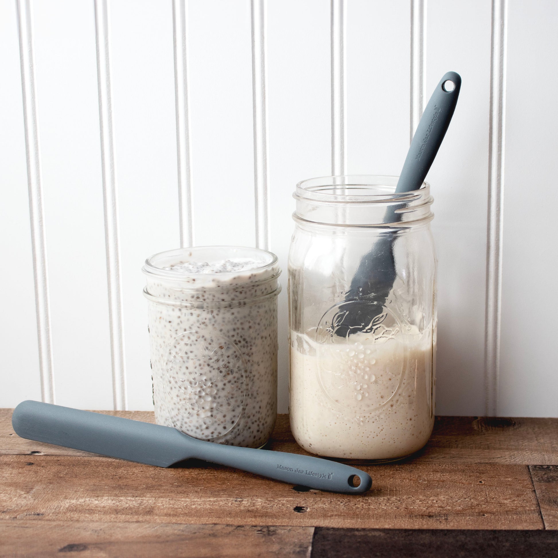 Two glass jars on a wooden surface—one with chia seed pudding, the other with a creamy liquid—showcase Mason Jar Lifestyle Silicone Jar Scrapers with ergonomic handles. A third scraper rests in front, all set against a white panel background.