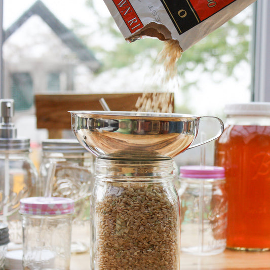 Brown rice is being poured through the Mason Jar Lifestyle Stainless Steel Canning Funnel for Mason Jars into a glass jar on a kitchen counter, surrounded by other containers, with a window in the background.