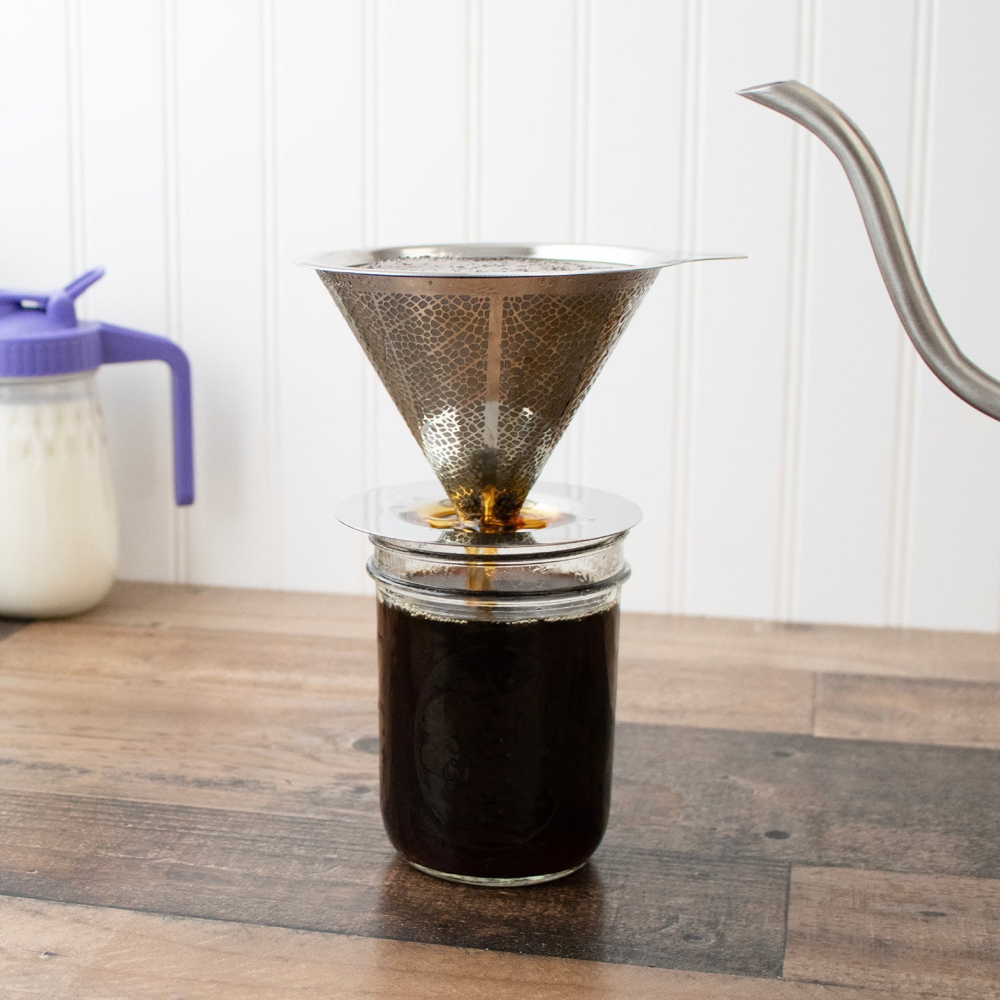 The Mason Jar Lifestyle Pour Over Drip Coffee Stainless Steel Filter rests on a Mason jar as coffee drips through, with a gooseneck kettle pouring hot water and milk in the background on a wooden counter.