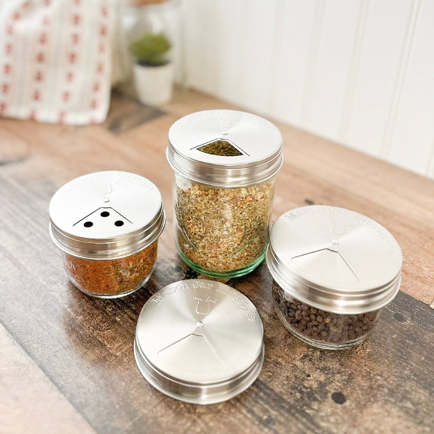 Mason Jar Lifestyle's Spice Shaker Stainless Steel Lids for Mason Jars 4 Pack are shown on a wooden countertop, each holding different spices or herbs. The adjustable lids allow for sprinkling, pouring, or scooping. A blurred plant is in the background.