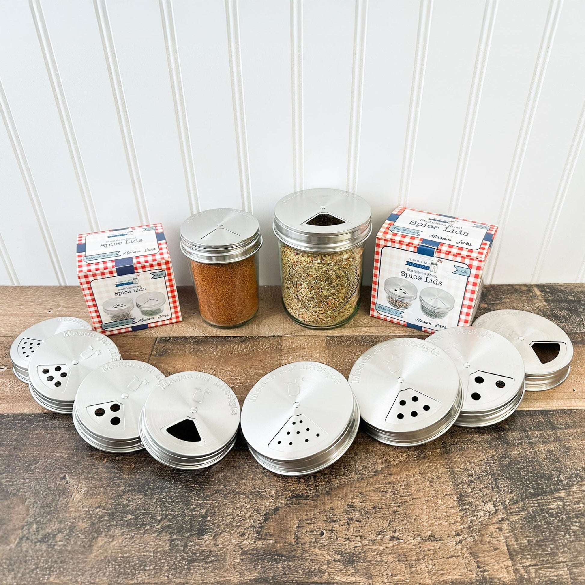 Mason Jar Lifestyle's Spice Shaker Stainless Steel Lids for Mason Jars (4 Pack) are displayed in a semicircle on wood, with two adjustable spice jars filled with spices and two red gingham boxes in the background.