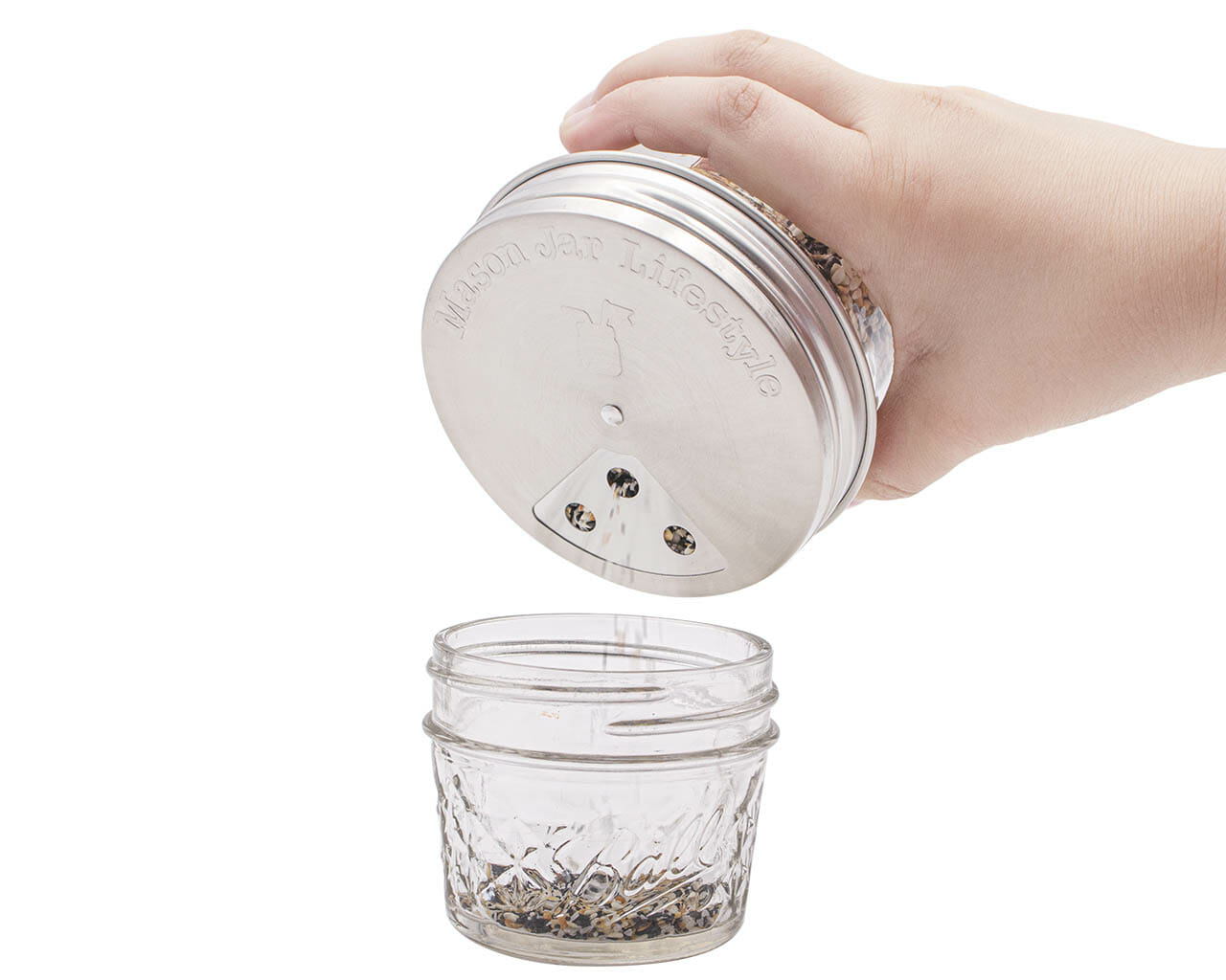A hand pours spices from a Mason Jar Lifestyle mason jar fitted with a Spice Shaker Stainless Steel Lid (4 Pack) into another empty mason jar, set against a white background.