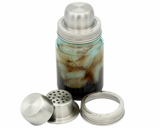 A Mason jar filled with ice and a dark liquid is topped with the Mason Jar Lifestyle Mason Shaker Cocktail Shaker Lid with Stainless Steel Band, with the shaker cap and strainer pieces shown in front of the jar.