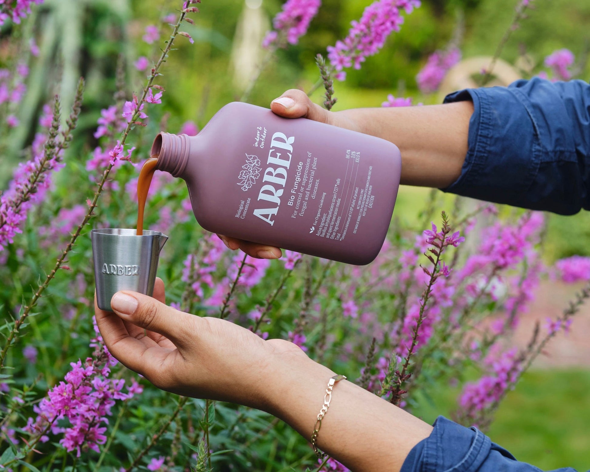 A person pours liquid from an Arber Measuring Cup into a stainless steel cup, surrounded by blooming purple flowers in a garden.